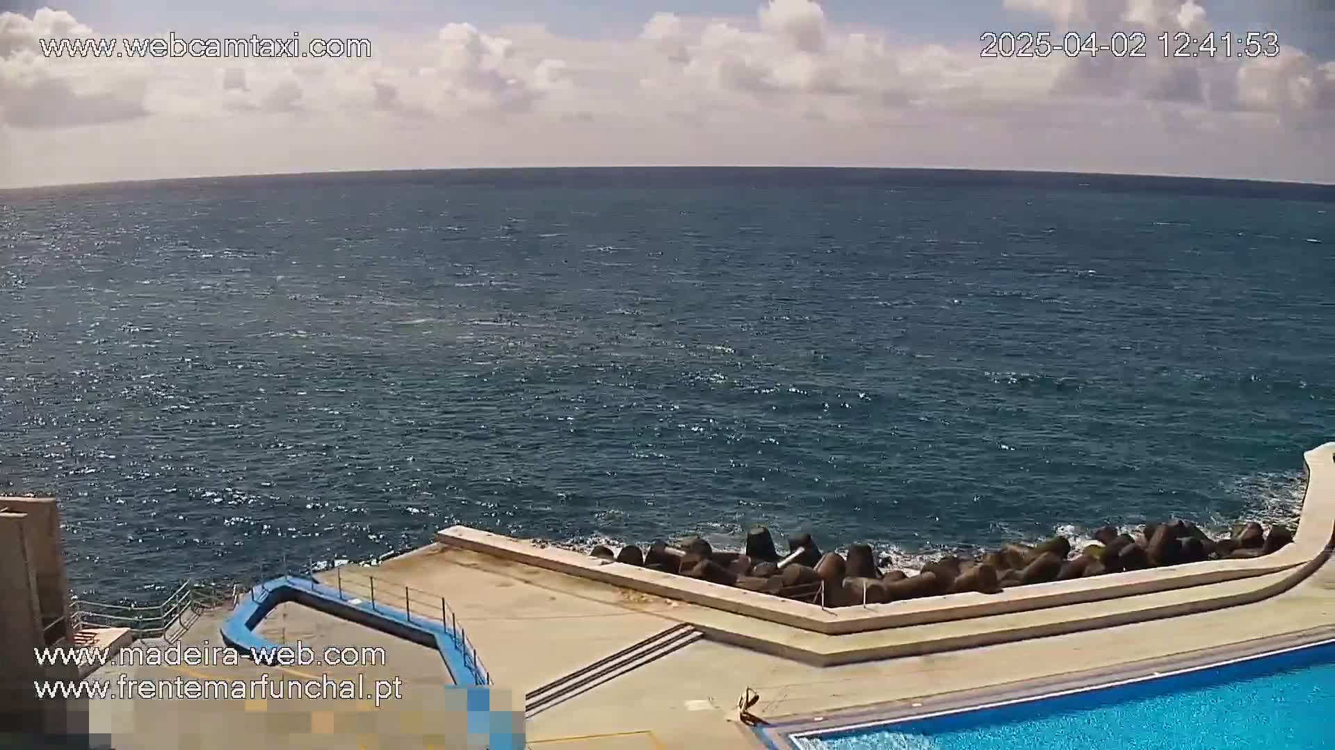 A partly cloudy day shows a blue ocean meeting a concrete structure with a pool adjacent to a breakwater.
