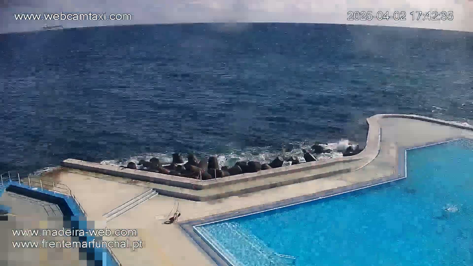 A partially overcast day shows a blue swimming pool adjacent to a rocky ocean shore with small waves crashing against a seawall.