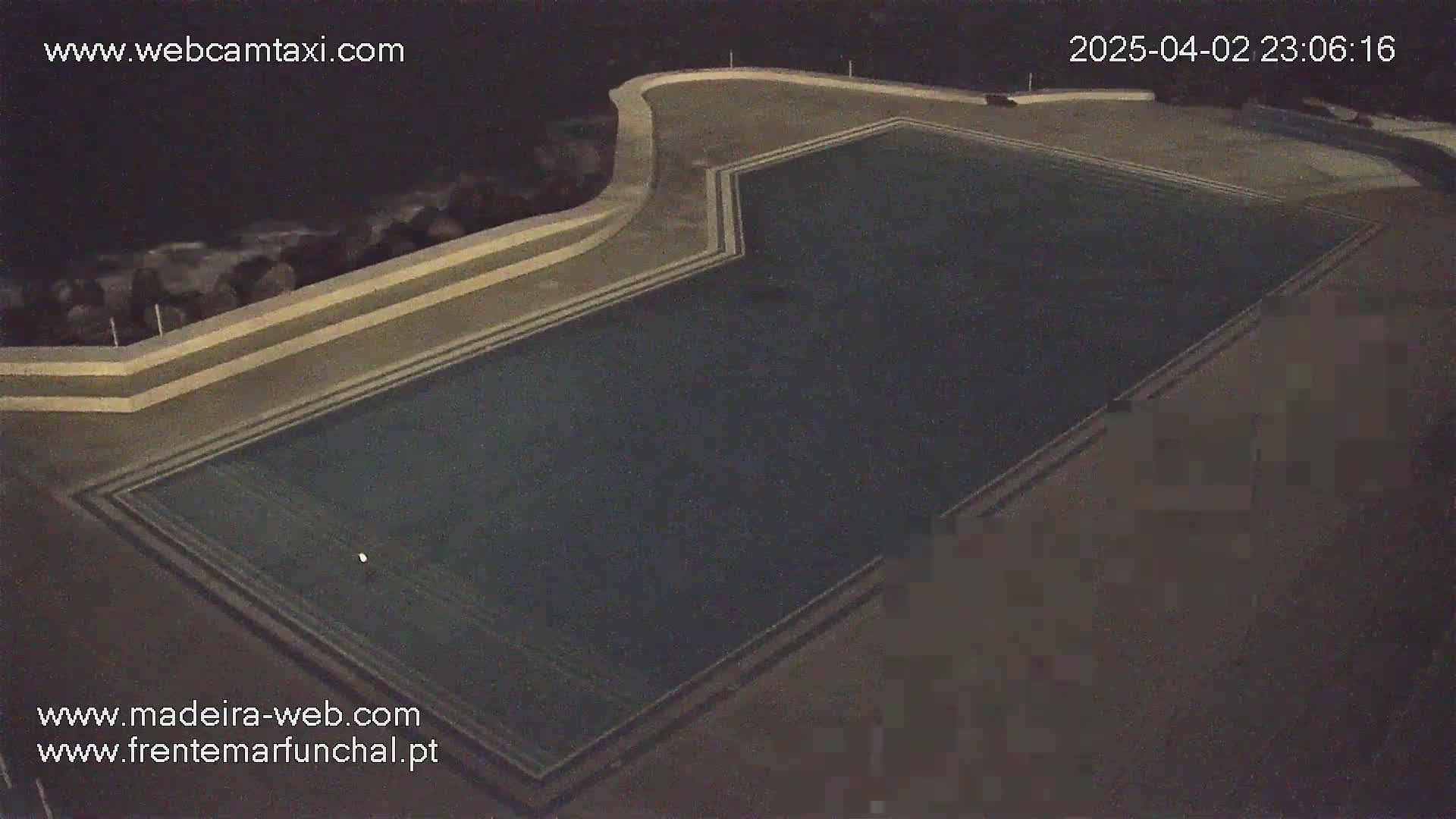 An outdoor nighttime aerial view shows a dark-colored rectangular swimming pool with white lines marking its lanes, surrounded by a dark gray concrete deck.