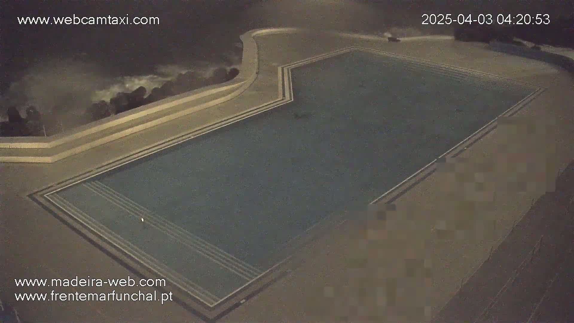 An empty swimming pool at night is shown, with rough ocean waves visible in the background.