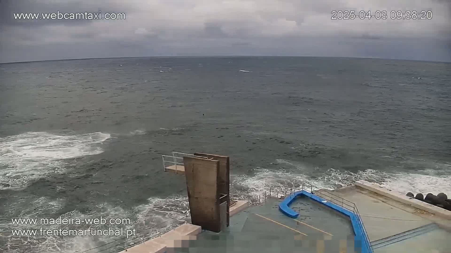 A high-angle view shows a choppy ocean under an overcast sky, with a diving platform and a partially visible pool area on the shore.