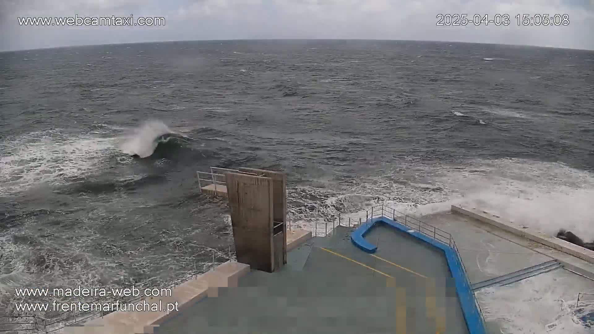 Rough ocean waves crash against a concrete structure with a small diving platform, under an overcast sky.