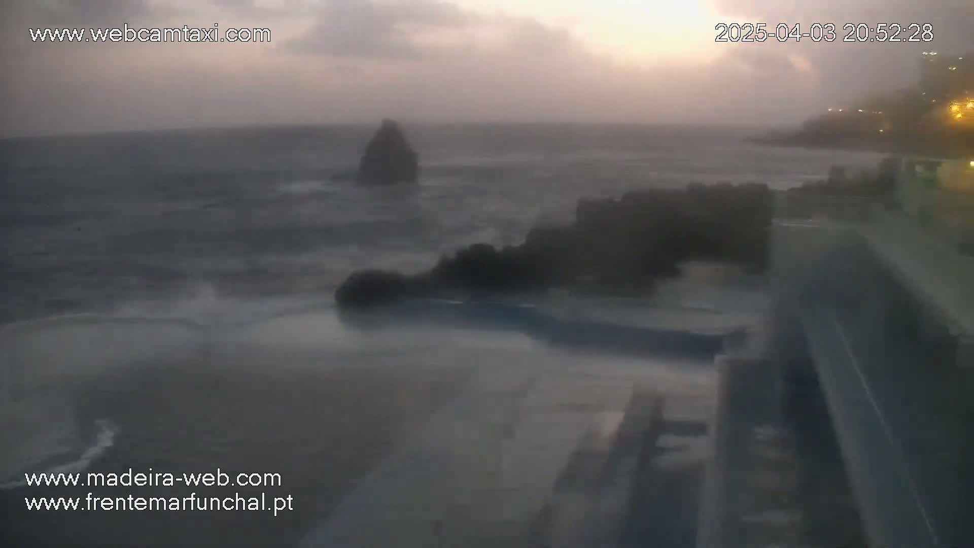 A stormy ocean scene at dusk shows dark, choppy waves crashing against rocky outcroppings near a shoreline with buildings.