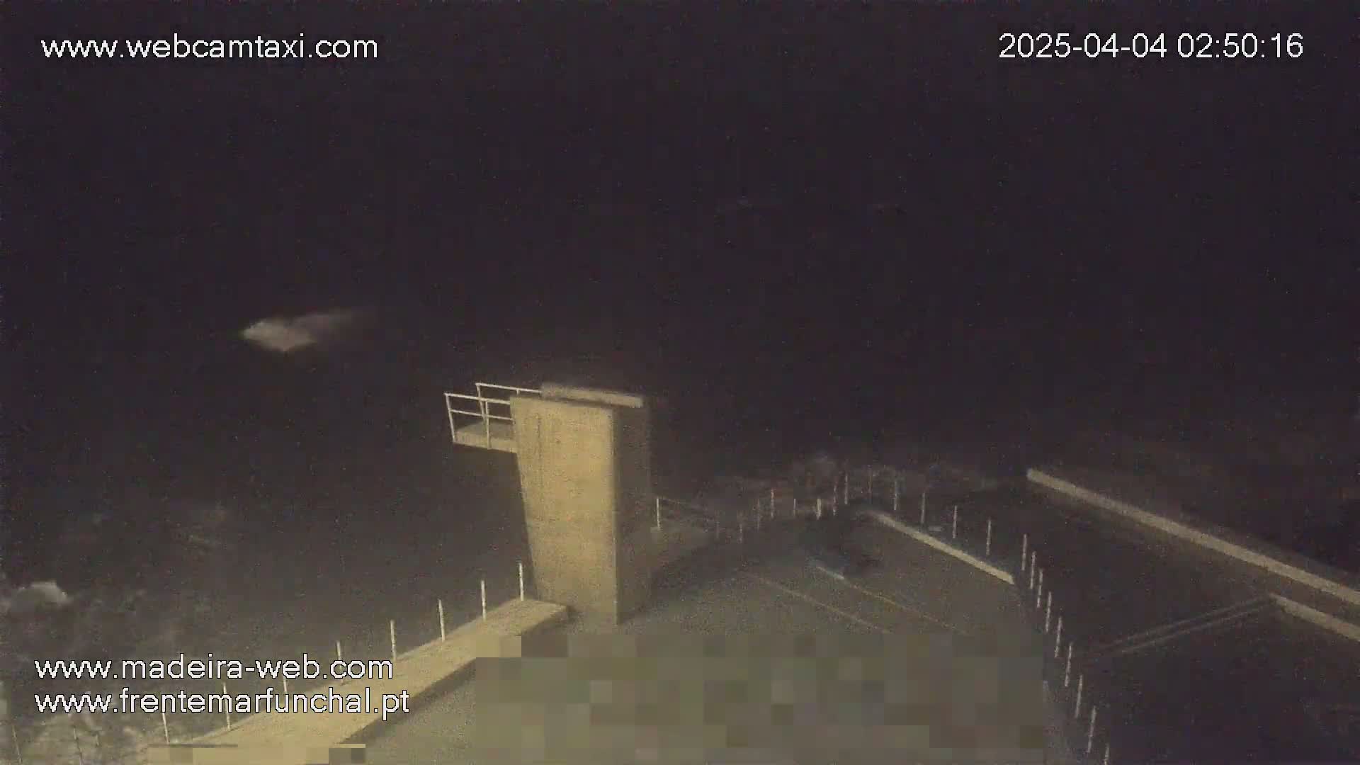 A nighttime, high-angle view shows a concrete structure resembling a diving platform and a parking lot, with a faint, light-colored object visible in the dark sky above.