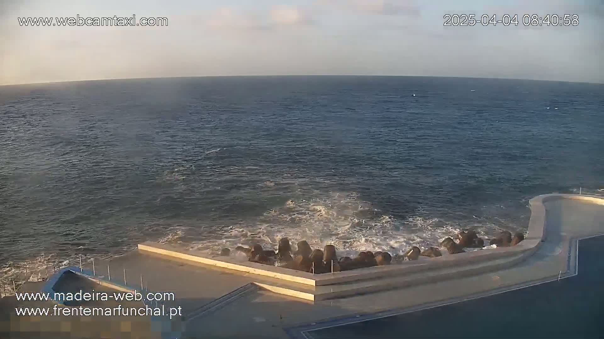 Ocean waves crash against a concrete structure on a calm, slightly hazy day.