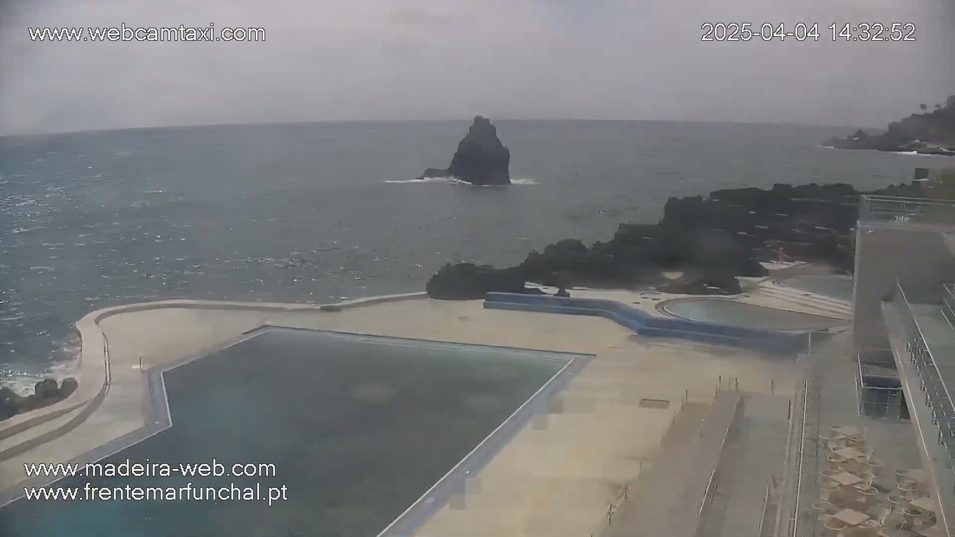 An outdoor scene shows a large, dark-colored pool next to a rocky coastline with choppy gray water and a small, dark island in the distance under an overcast sky.