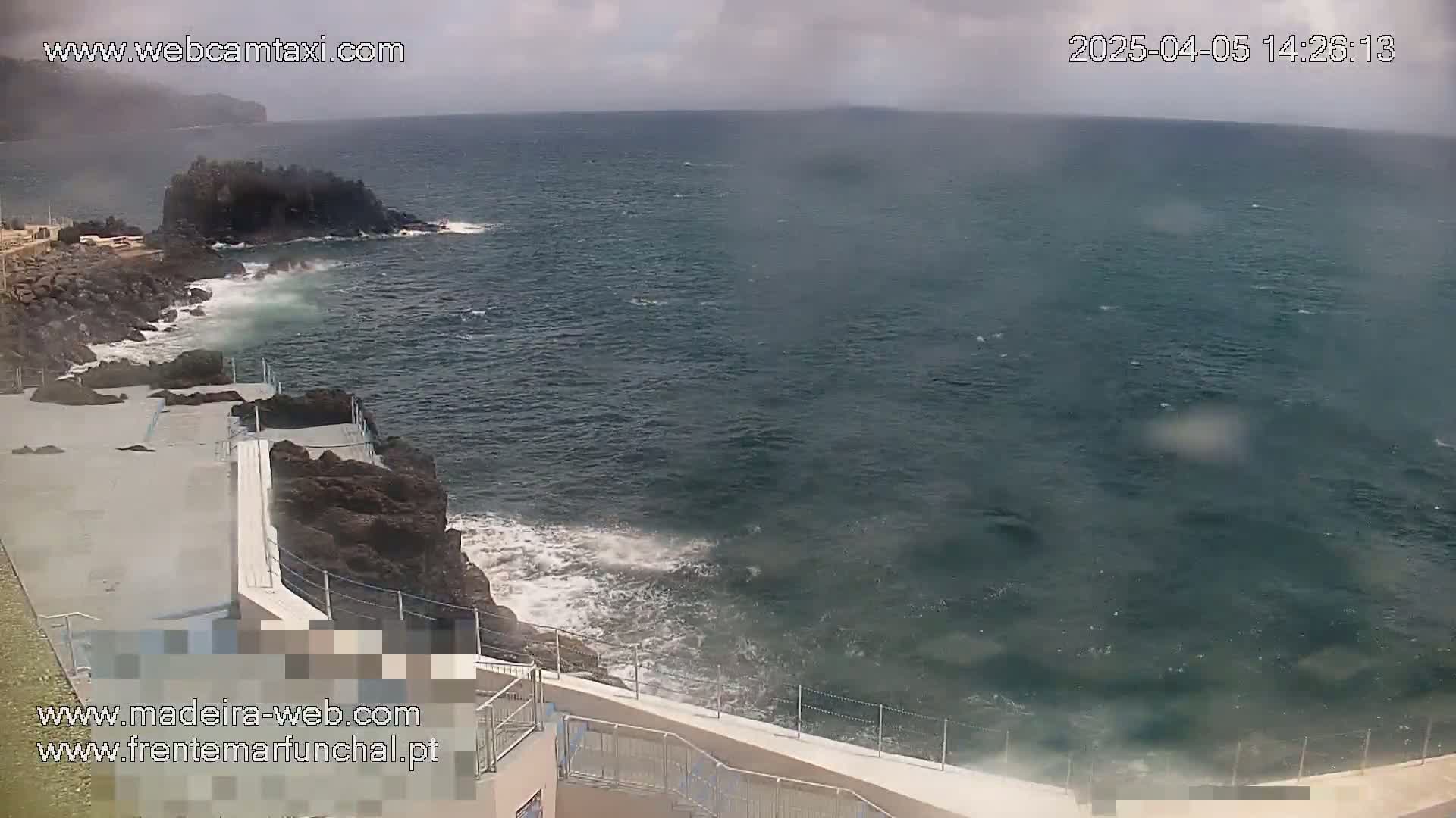 A rocky coastline with a concrete walkway and railings overlooks a dark-blue ocean with moderate waves under an overcast sky.