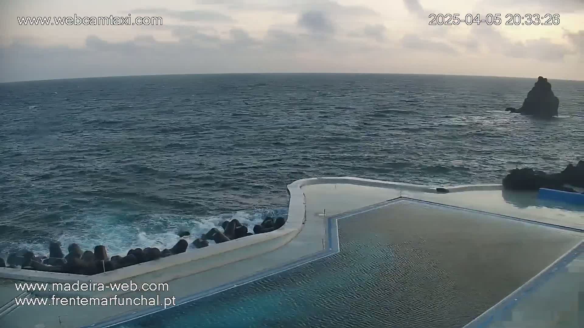An infinity pool overlooking a dark-blue ocean with small waves crashing against a seawall under an overcast sky.