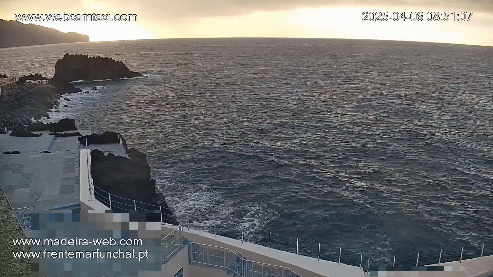 A rocky coast with a paved area and railing overlooks a dark, somewhat choppy ocean under an overcast sky.