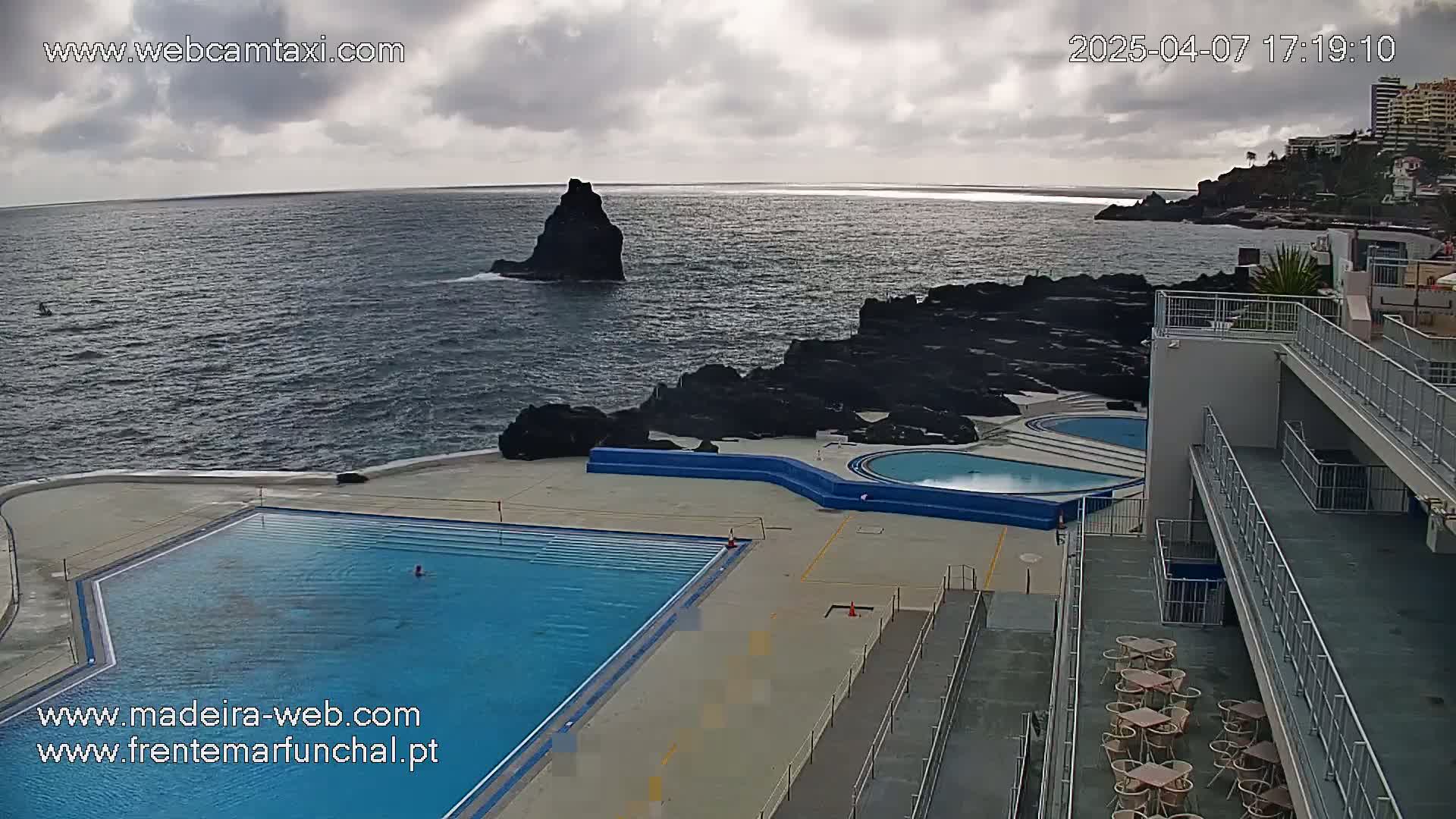 An outdoor swimming pool complex sits on a rocky coastline overlooking a dark blue ocean under a cloudy sky.