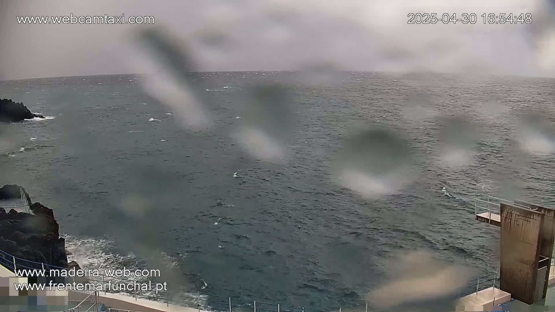 A choppy, gray sea is viewed from a rocky shore on an overcast day, with rain obscuring the view and a diving platform visible in the lower right corner.