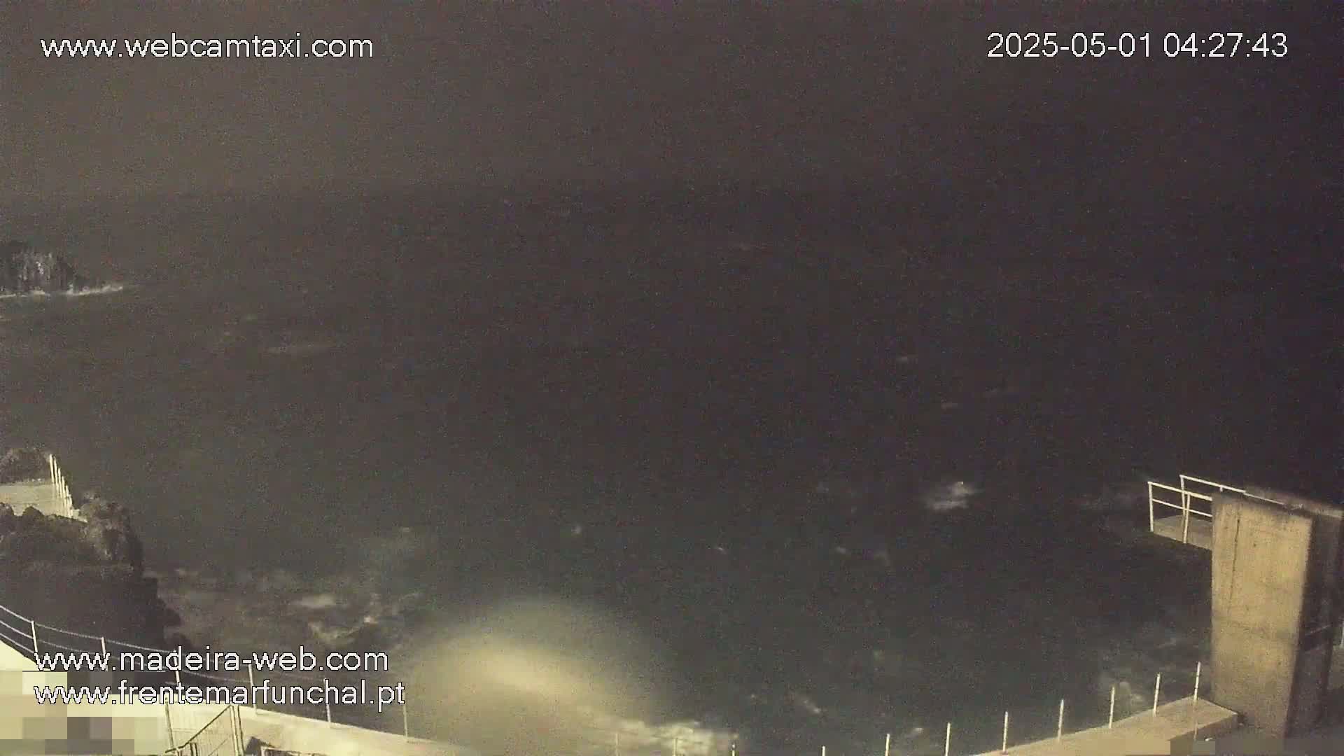 A nighttime view shows dark, choppy water crashing against a rocky shore near a low concrete structure.