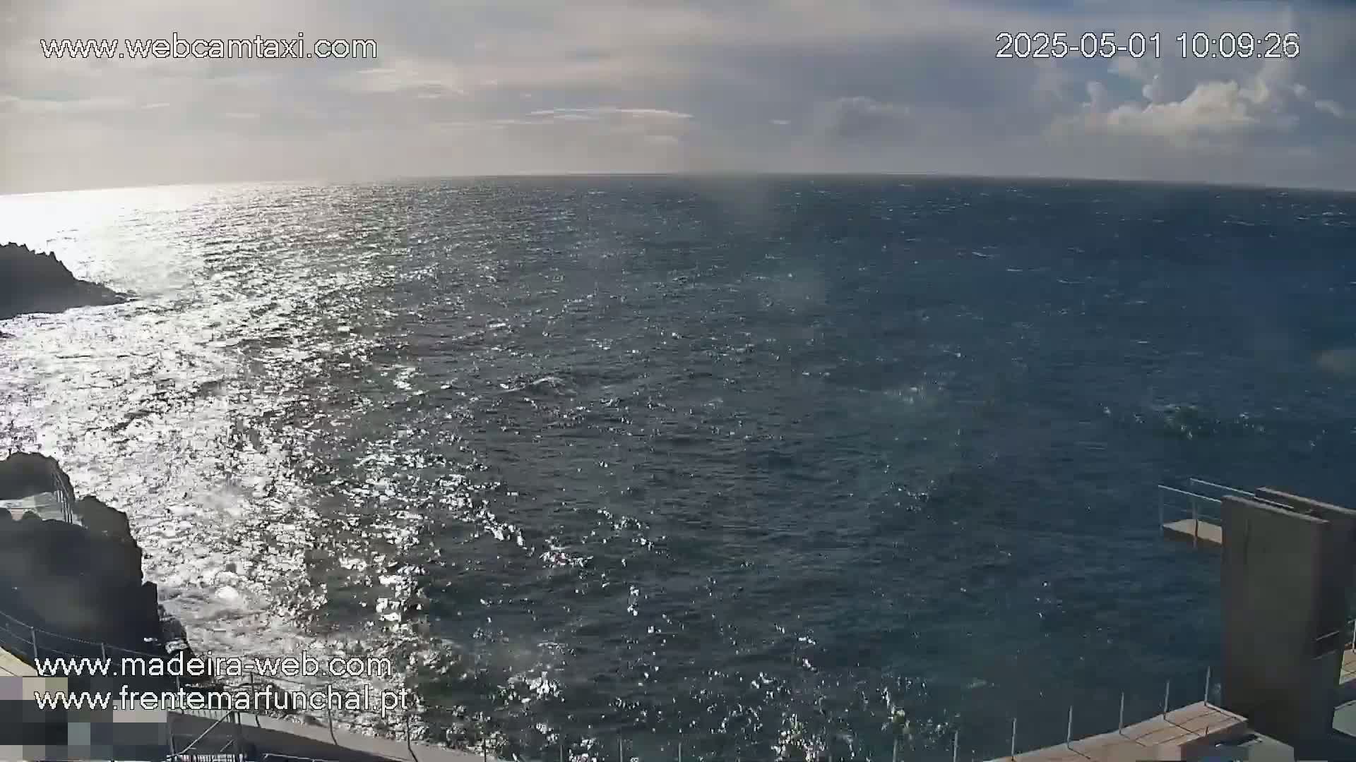 A somewhat choppy ocean with a partly cloudy sky is viewed from a high vantage point, showing dark-colored rocks and a low concrete wall at the bottom of the frame.