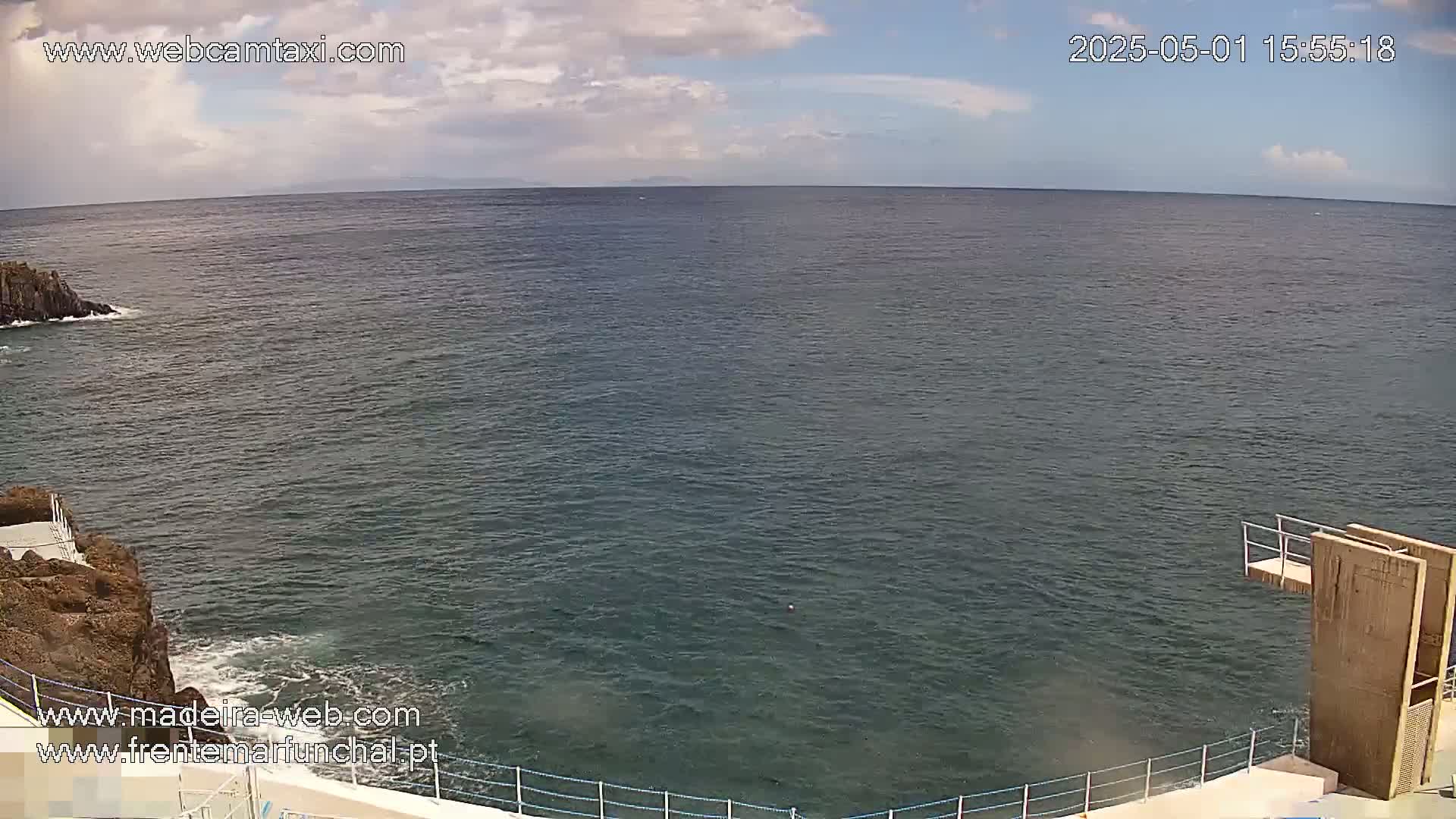 A mostly cloudy sky overlooks a dark-blue ocean meeting dark-colored rocky shores with a diving platform at the bottom right.