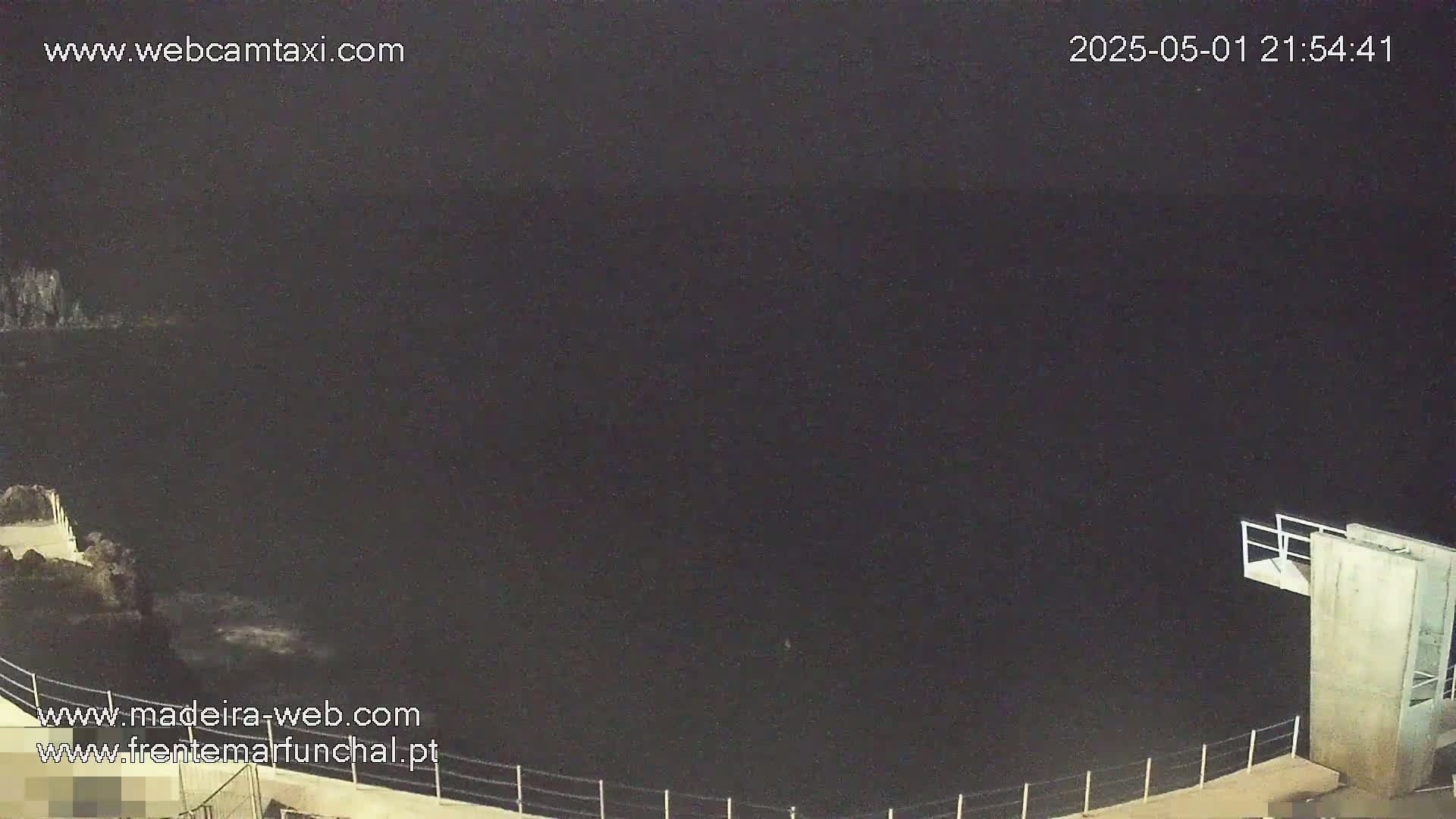 A dark, nighttime view of a body of water from an elevated vantage point, showing a concrete structure with a diving platform extending over the water's edge.