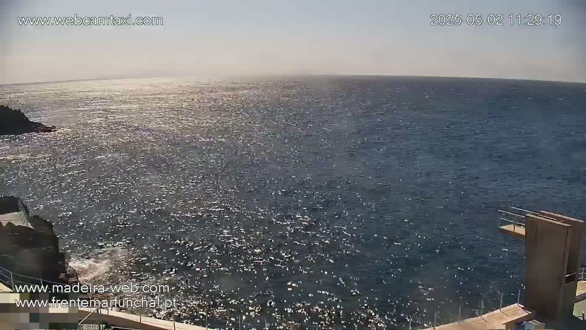 A sunny, bright day shows a vast, sparkling ocean meeting a rocky shore with a diving platform.