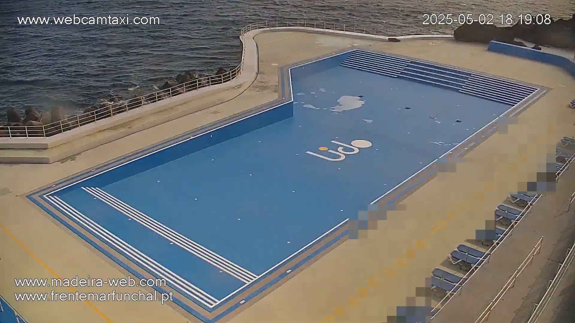 An outdoor, rectangular swimming pool with multiple sets of steps at one end sits next to a rocky ocean shore, with several empty lounge chairs along the edge of the pool deck.