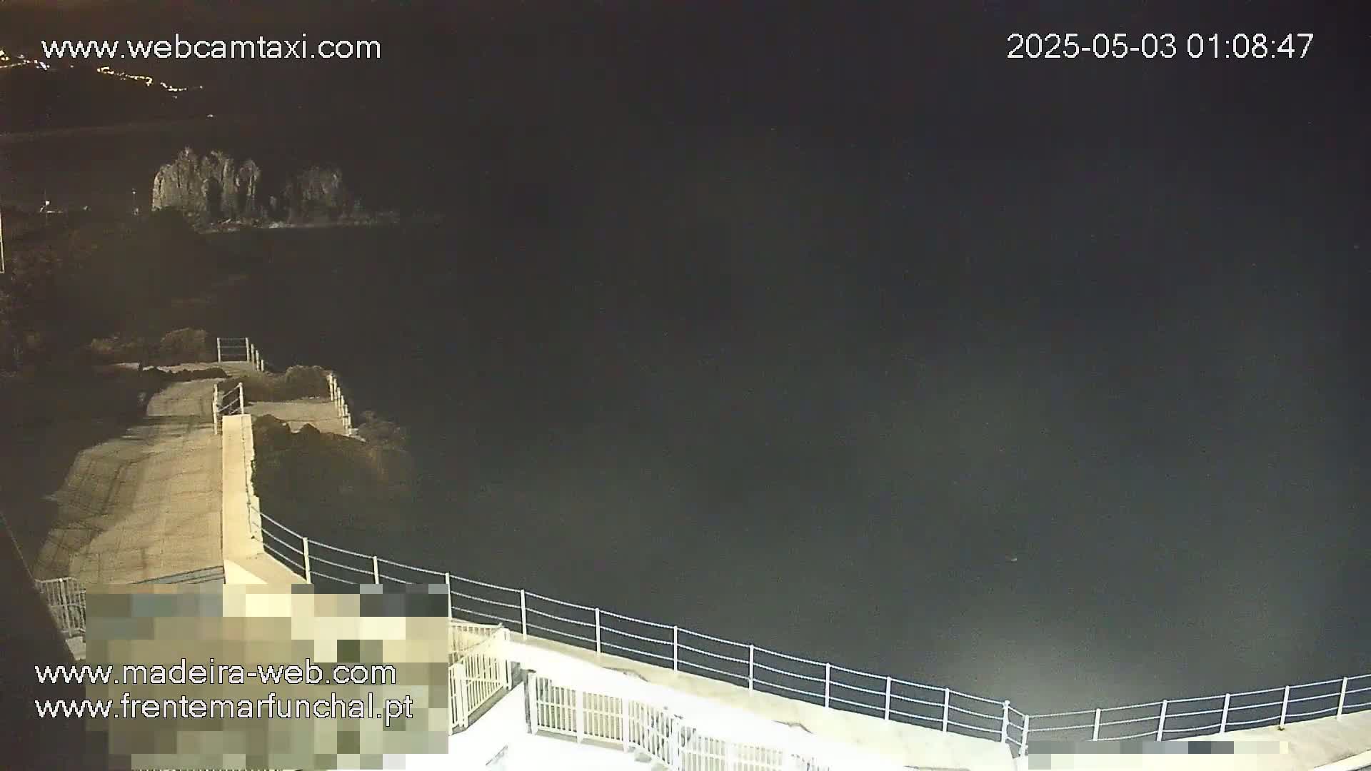 A nighttime, high-angle view shows a paved walkway along a dark body of water, bordered by a low metal railing.