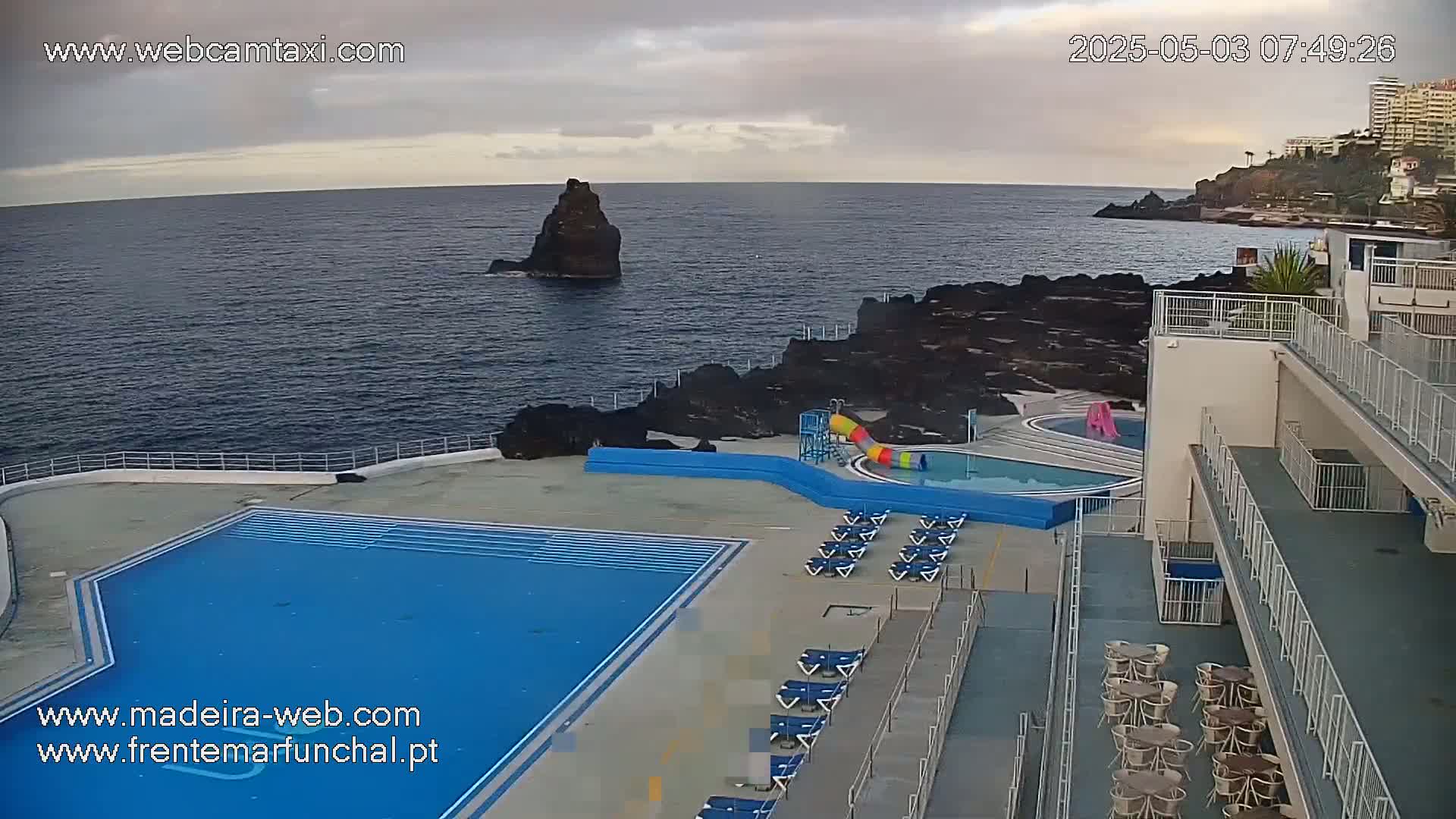 An outdoor pool and surrounding patio area with lounge chairs and tables overlook a dark blue ocean and rocky coastline under a partly cloudy sky.