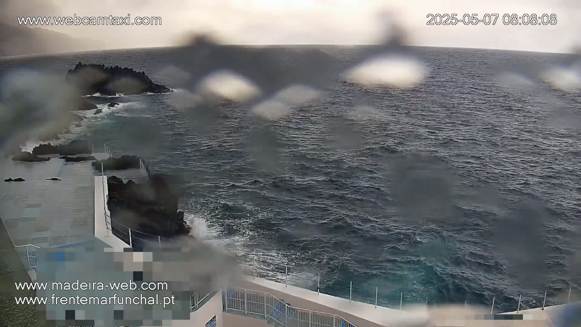 A rain-spattered view shows dark, choppy ocean waves crashing against dark rocks near a light-colored concrete structure.