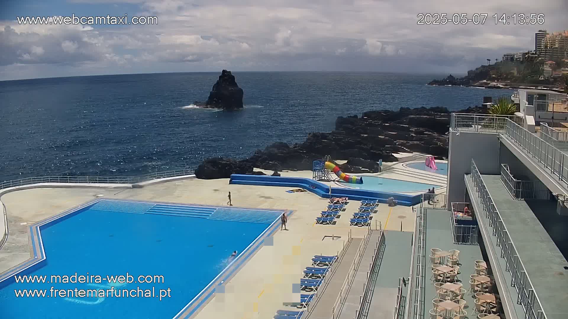 An outdoor pool complex with a smaller children's pool and water slide is situated on a rocky coast overlooking a dark blue ocean under a partly cloudy sky.