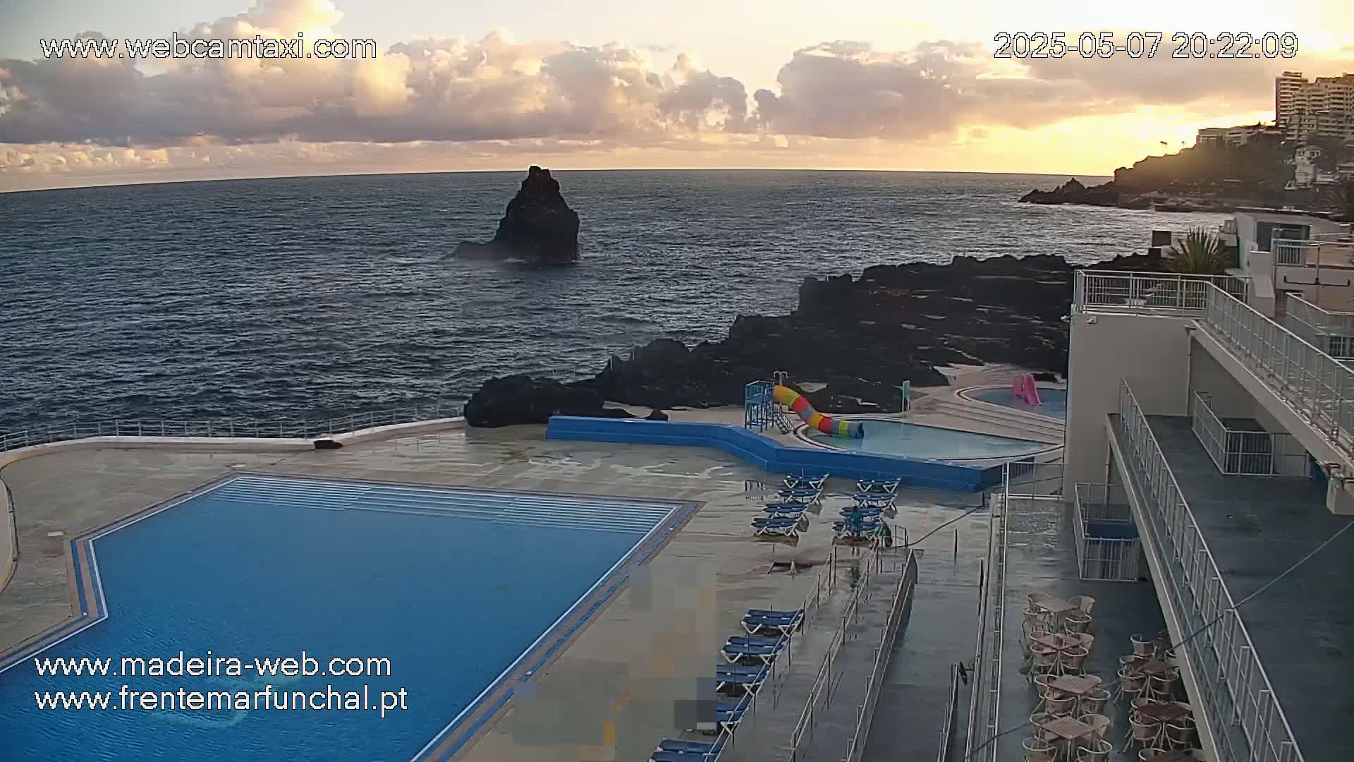 An outdoor swimming pool complex with a waterslide sits on a rocky coast overlooking a dark blue ocean under a partly cloudy sunset sky.