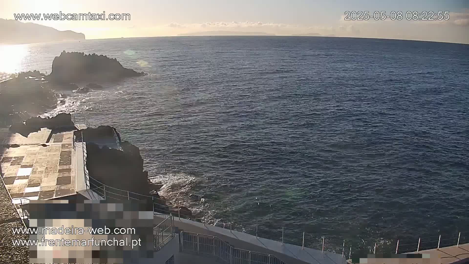 A rocky coastline with a paved area and railings overlooks a calm ocean under a mostly clear sky.