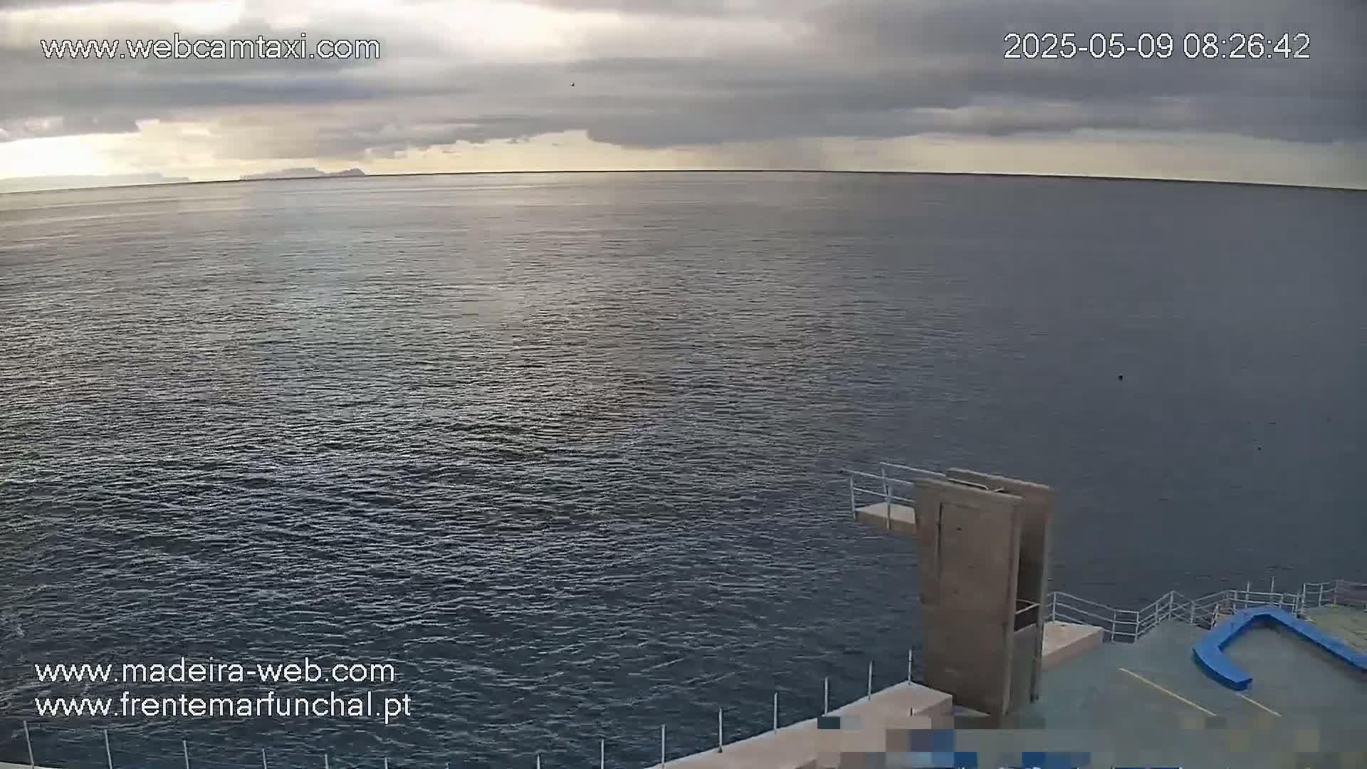 A wide shot of a calm, dark-blue ocean under a cloudy sky shows a diving platform extending into the water.