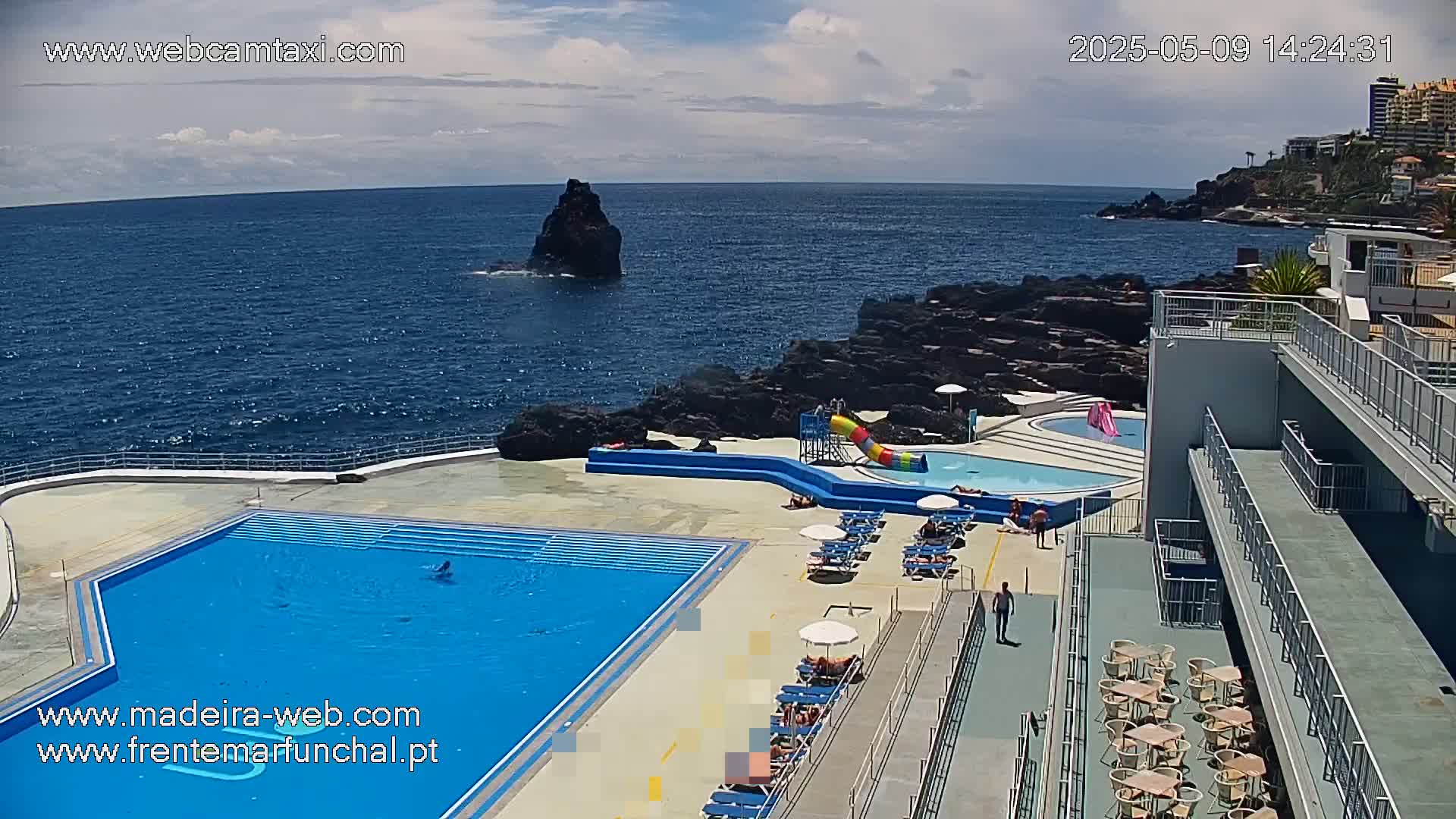 An outdoor pool complex with a large swimming pool and a smaller pool with a water slide overlooks a dark blue ocean on a partly cloudy day.