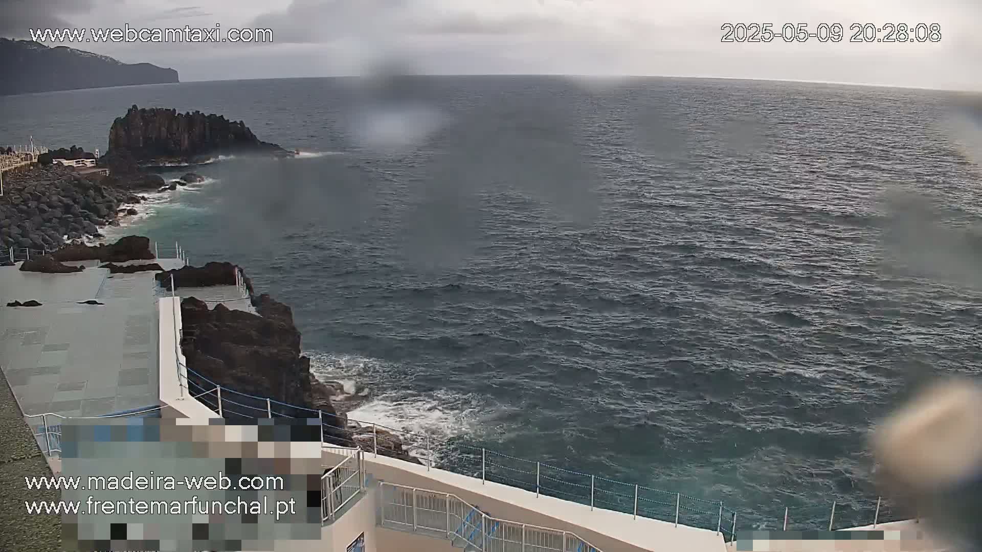 A rocky coastline with a concrete structure overlooks a dark, choppy ocean under an overcast sky.