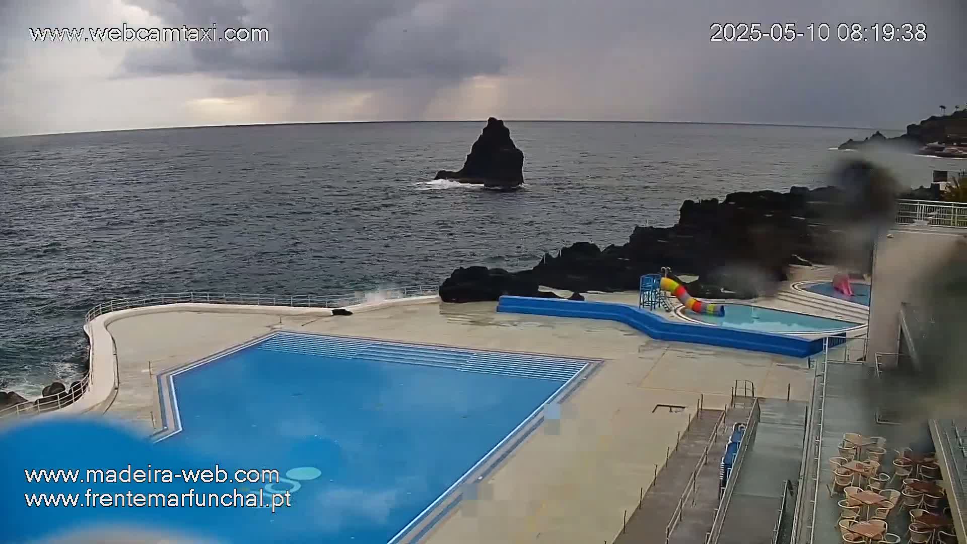 An outdoor pool complex sits on a rocky coast overlooking a dark, choppy ocean under a cloudy sky.