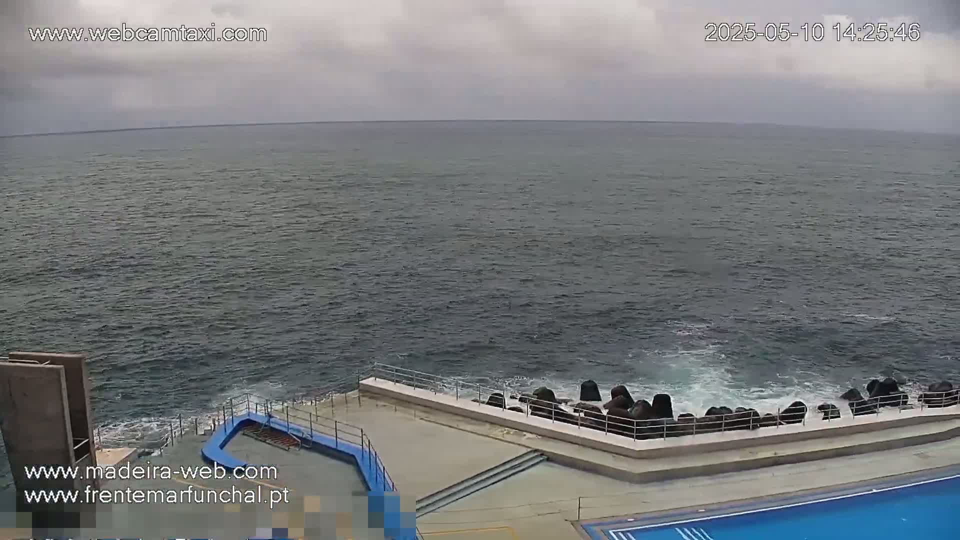 A mostly cloudy day shows a view of a dark-blue ocean meeting a light-grey concrete structure with a partially visible swimming pool, all bordered by dark rocks.