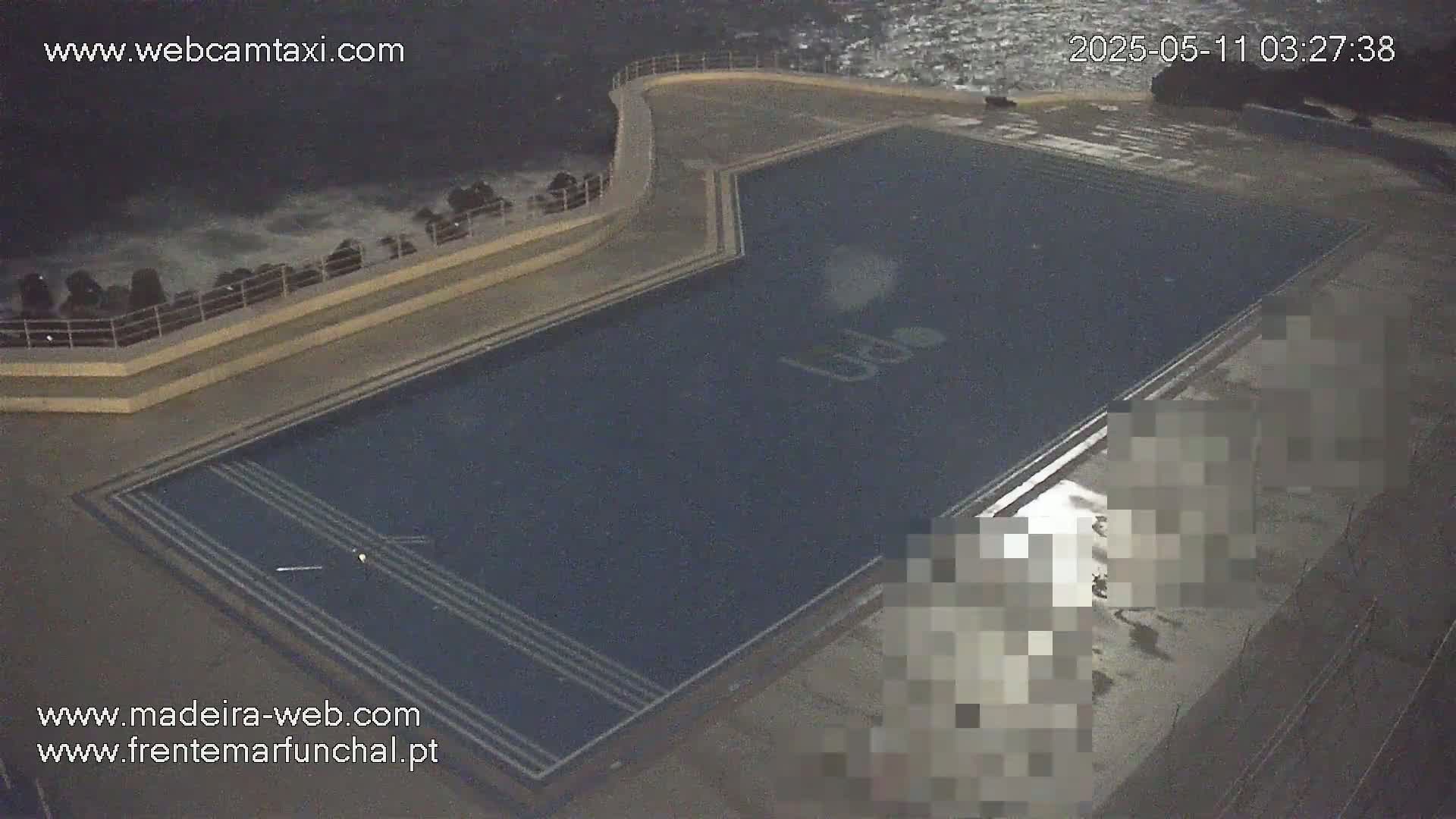 An empty, dark-blue outdoor pool sits next to a rocky coast with rough waves at night.