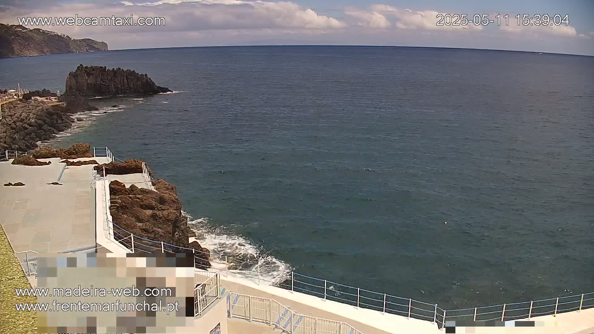 A partly cloudy day shows a rocky coastline with a white concrete walkway and railing overlooking a dark blue ocean with small waves.