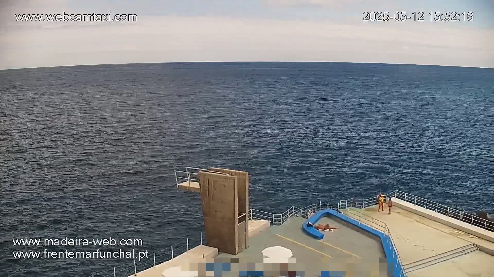 A diving platform overlooks a calm, dark blue ocean under a mostly cloudy sky; two people stand near the platform, and another person sunbathes on the concrete beside it.