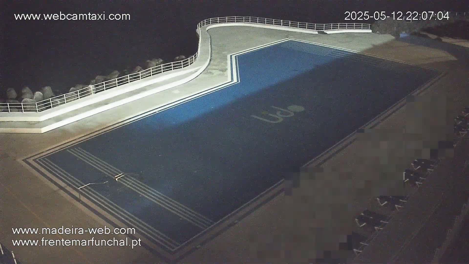 An outdoor, nighttime view of a dark-blue rectangular swimming pool with white lines and a logo, situated alongside a white concrete wall and railing overlooking a dark body of water.