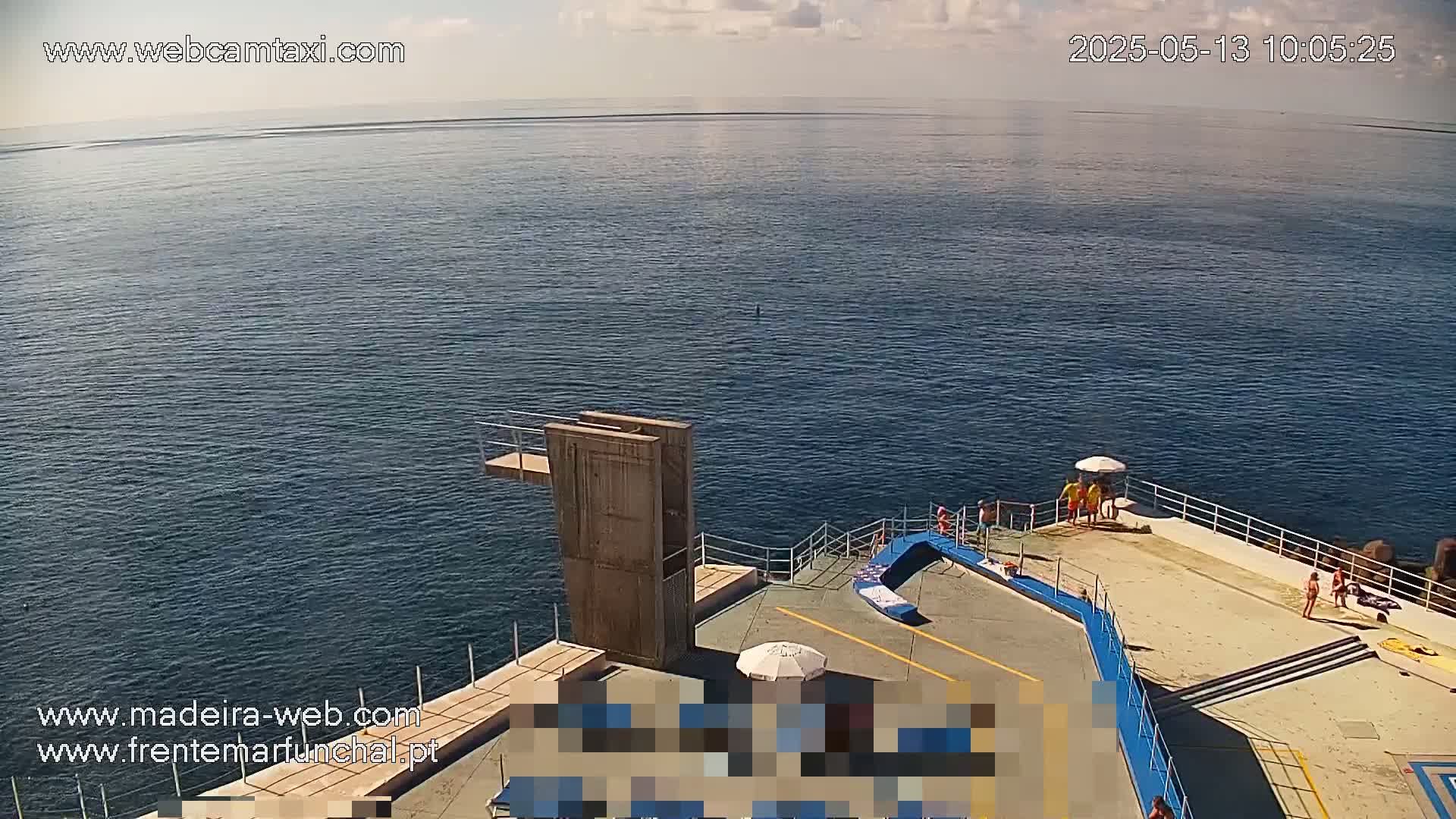 A mostly sunny day overlooking a calm ocean with a diving platform and several people on a concrete platform near the water.