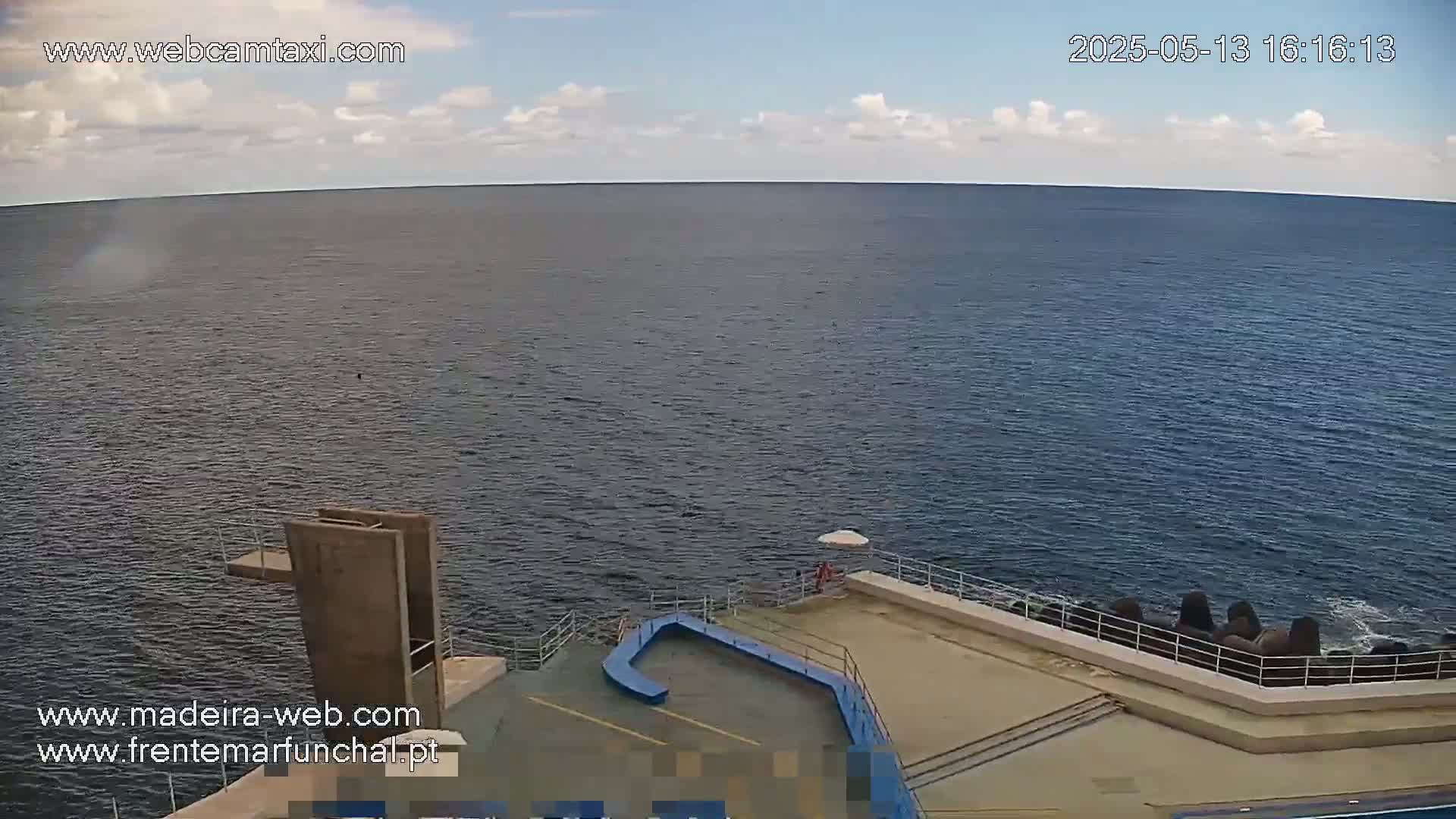 A mostly sunny day overlooking a calm ocean, showing a concrete structure with a diving board near a breakwater.