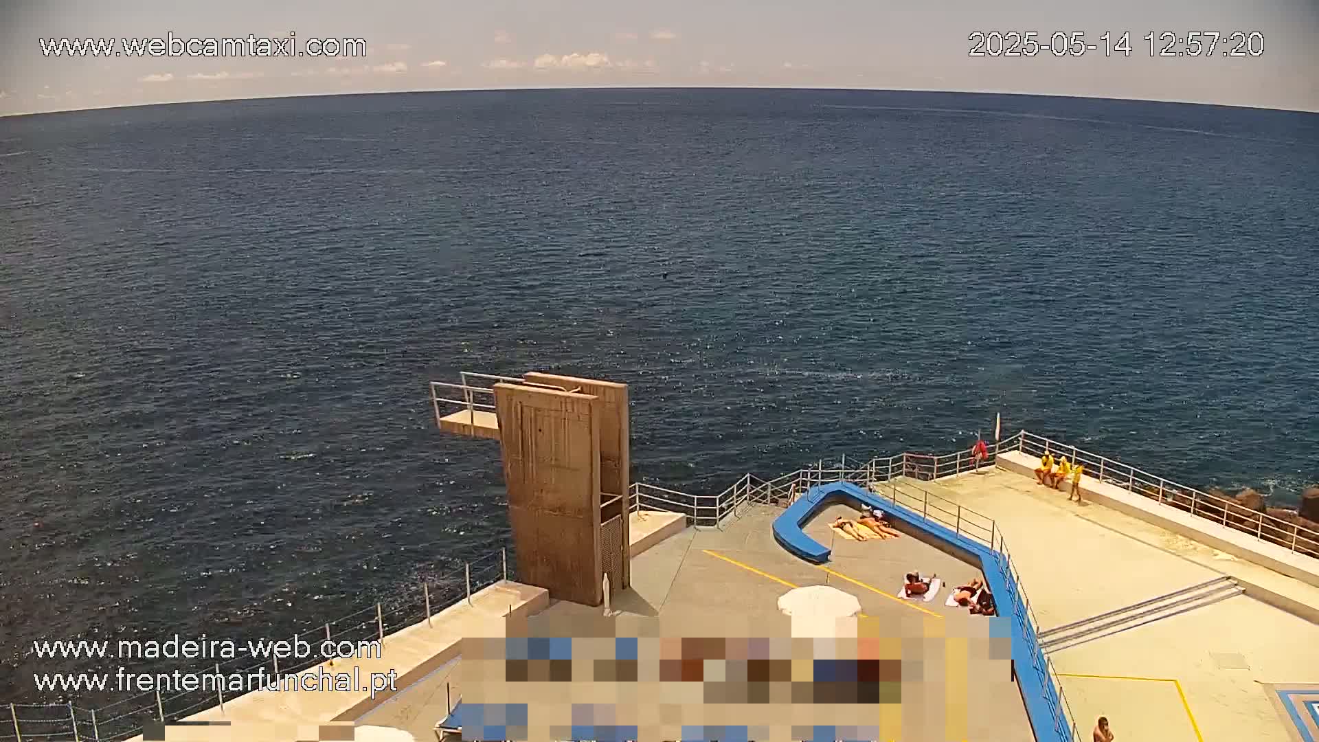 A high-angle, sunny view shows a concrete seaside platform with sunbathers and lifeguards, a diving board, and dark blue ocean extending to the horizon.