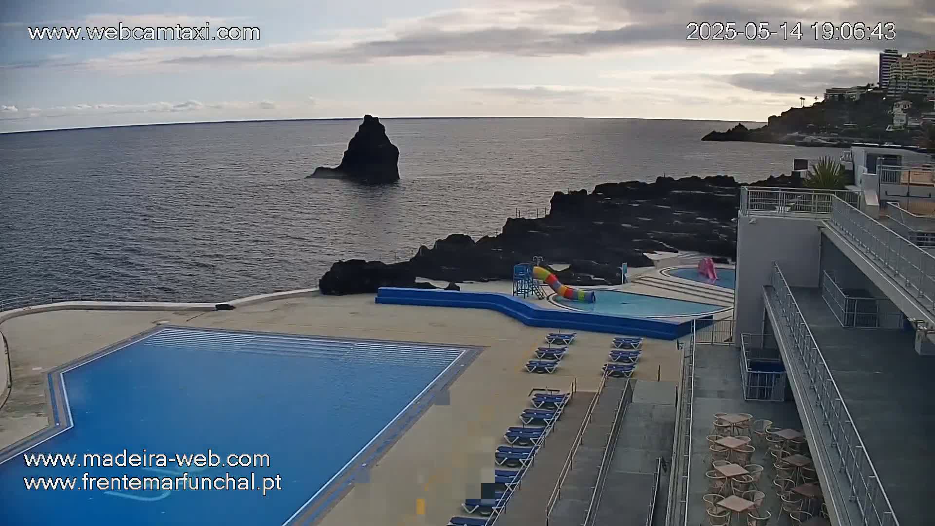 An outdoor pool area with lounge chairs and a water slide overlooks a dark-blue ocean under a cloudy sky.