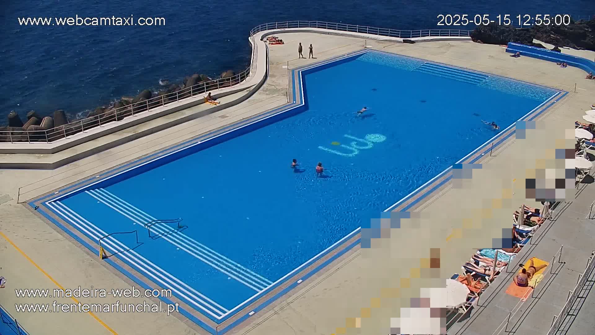 A large outdoor pool, mostly blue, is situated next to the ocean on a sunny day, with several people swimming and sunbathing nearby.