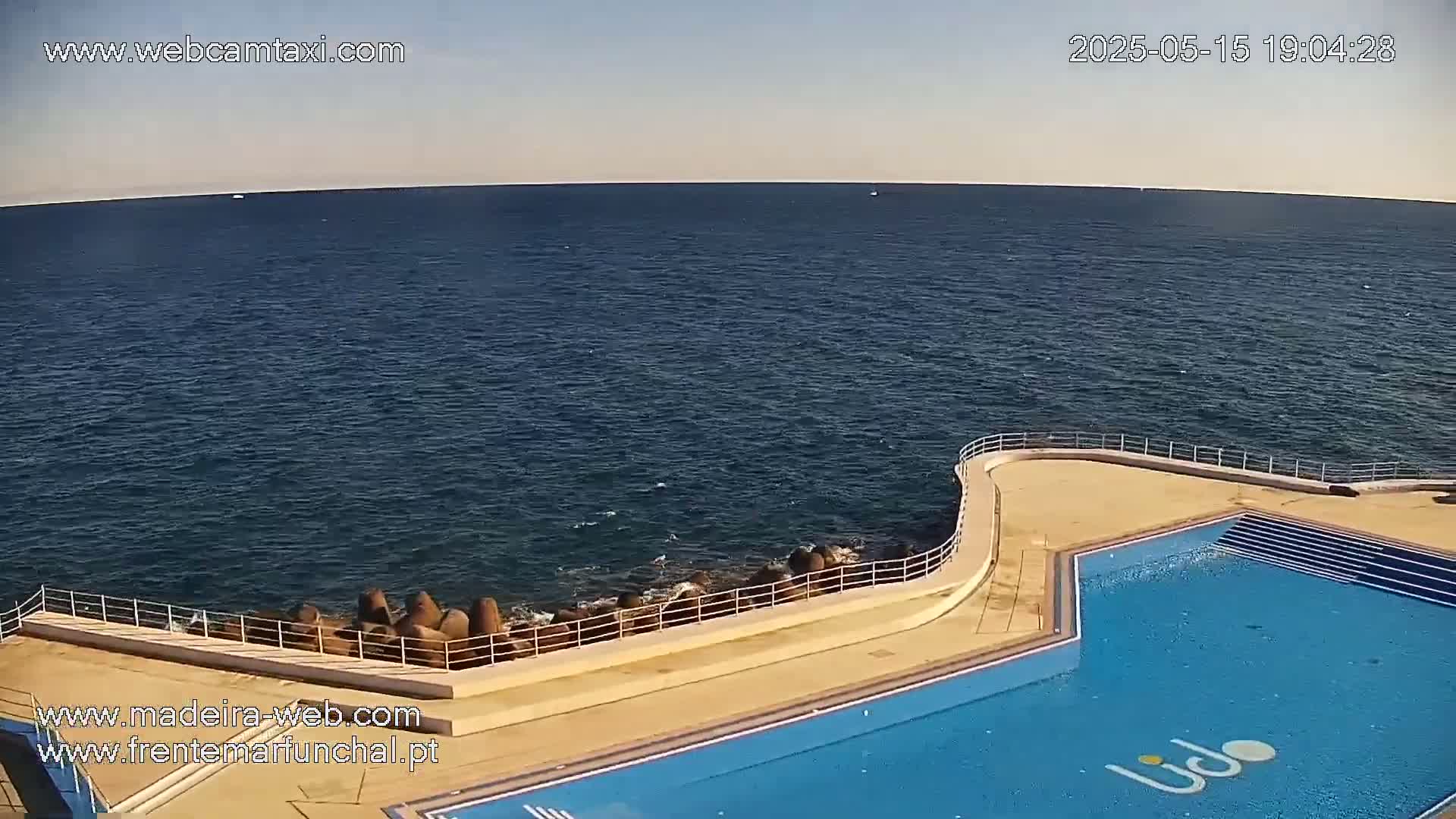 An outdoor swimming pool is situated on a concrete platform overlooking a calm, dark blue ocean under a clear sky.