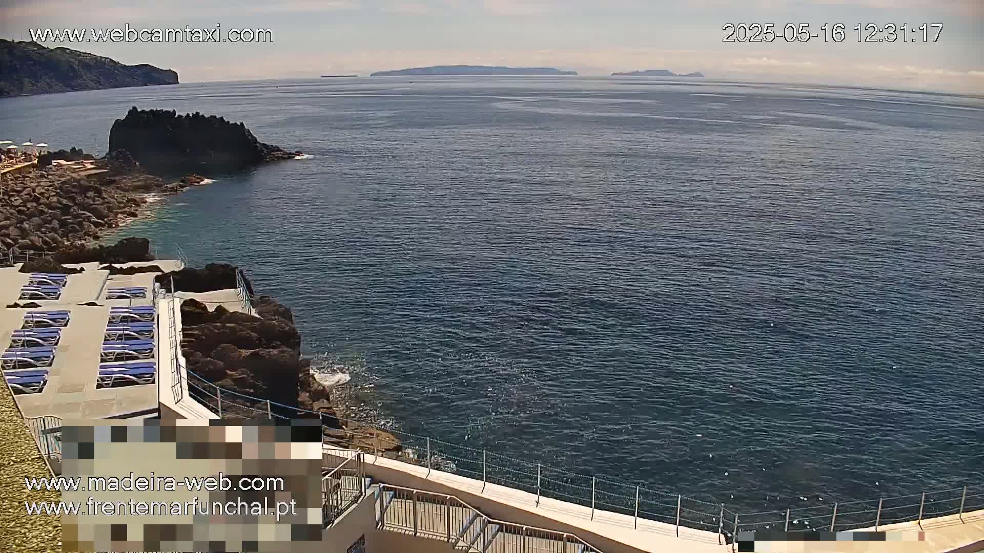A rocky coastline with a sun deck and lounge chairs overlooks a calm ocean under a mostly clear sky.
