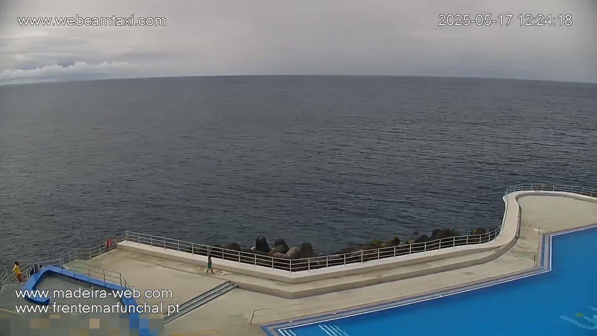 An overcast day shows a view of a dark-blue ocean with a concrete pool and patio area next to it, featuring a low wall and a few people.
