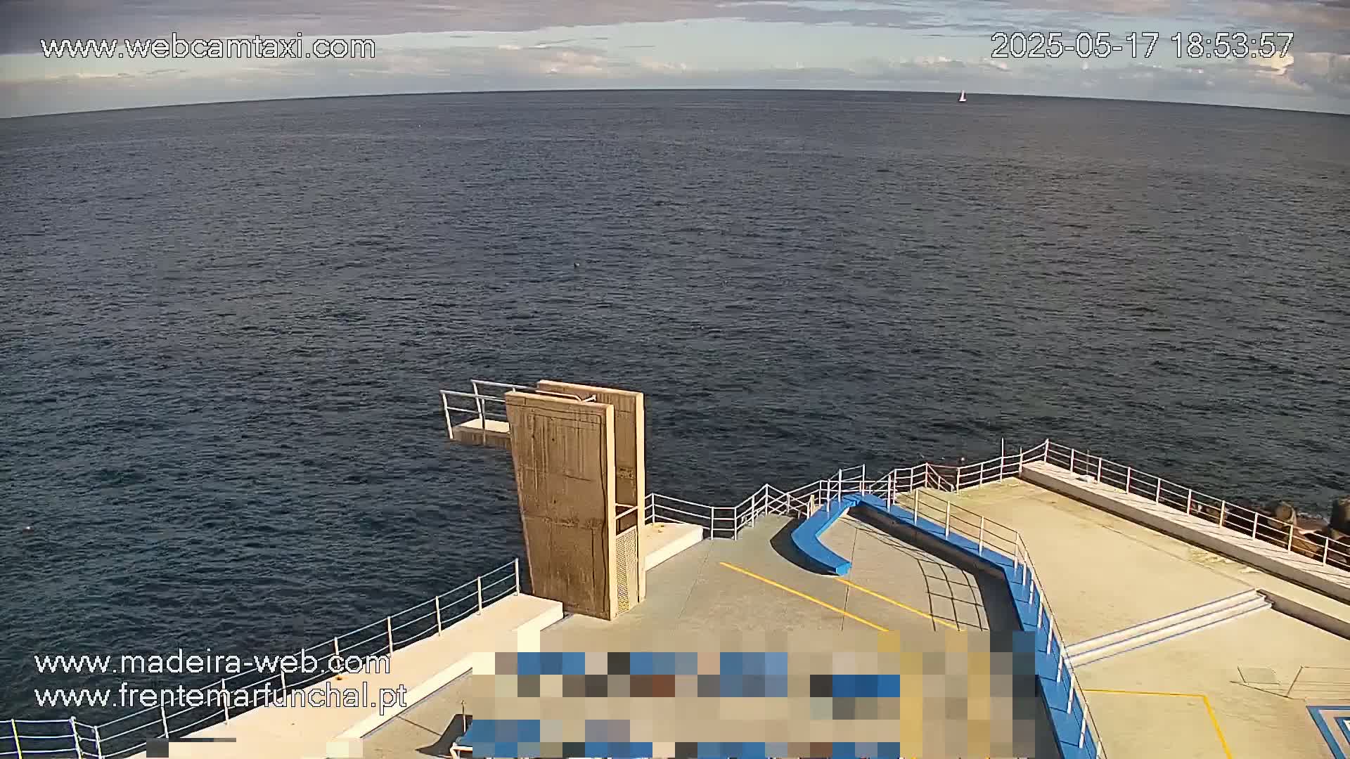 A high-angle view shows a diving platform extending over a calm, dark-blue ocean under a partly cloudy sky.
