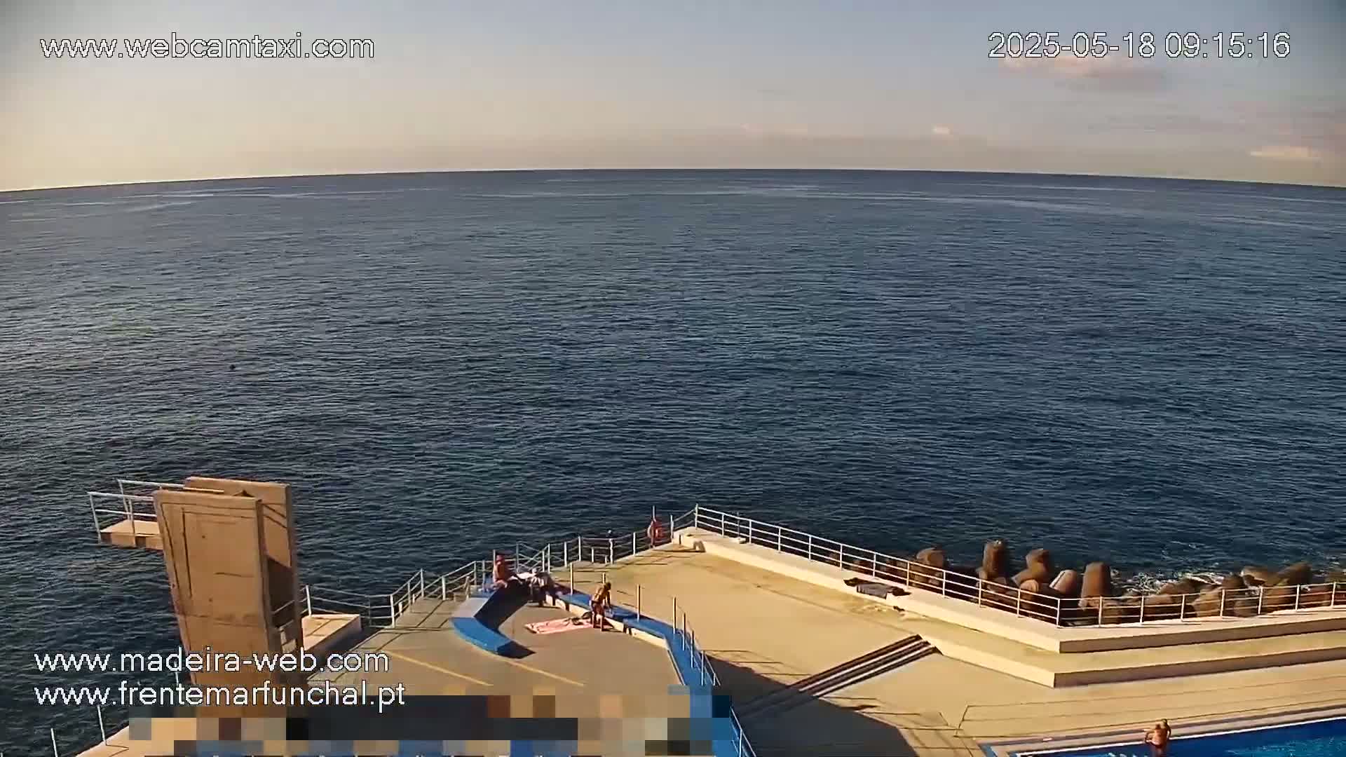A calm ocean is visible from an elevated vantage point overlooking a concrete platform with a diving board and a few people sunbathing near a small pool.