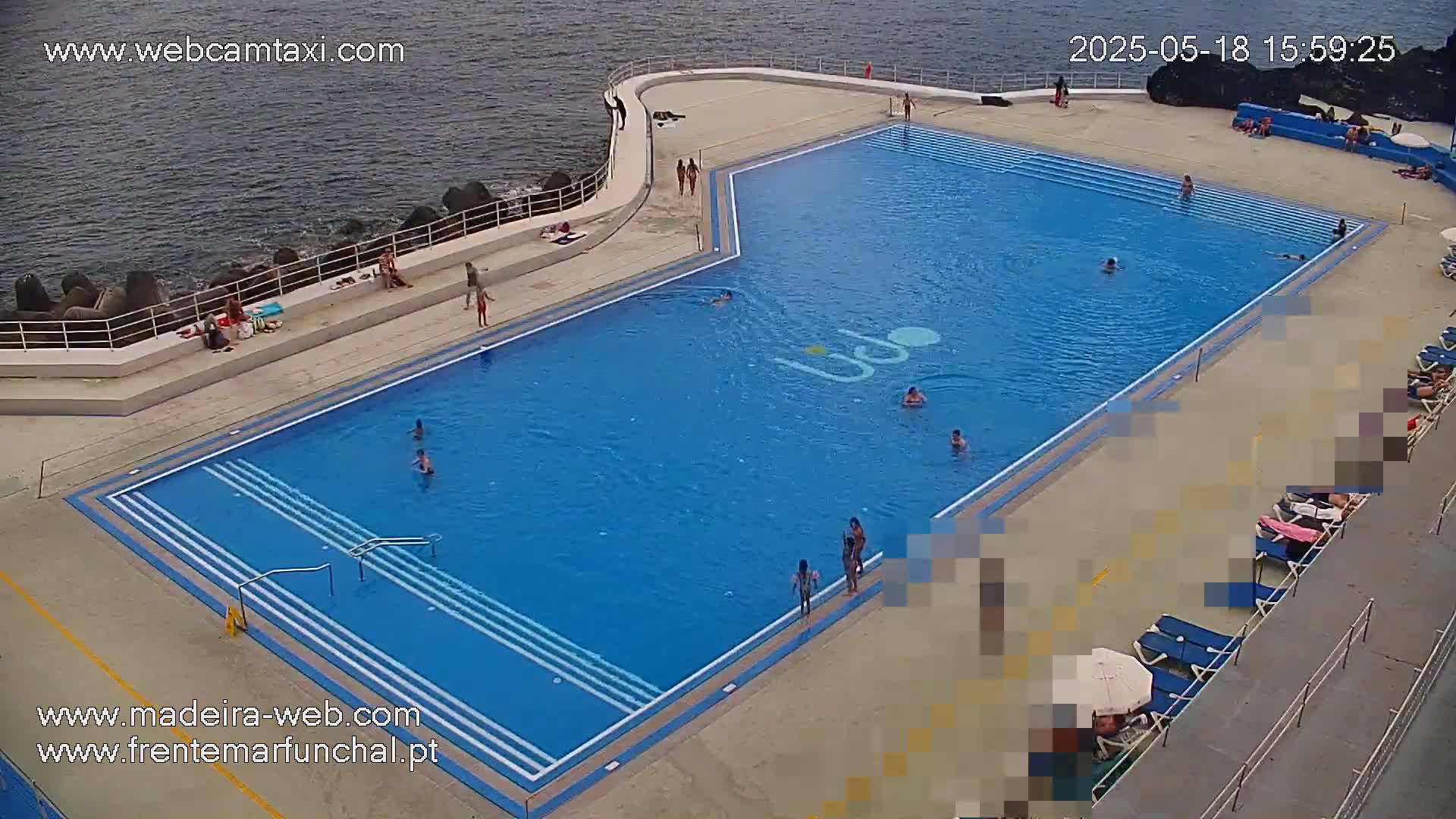 An outdoor swimming pool, mostly filled with water, is surrounded by people relaxing and swimming on a sunny day.