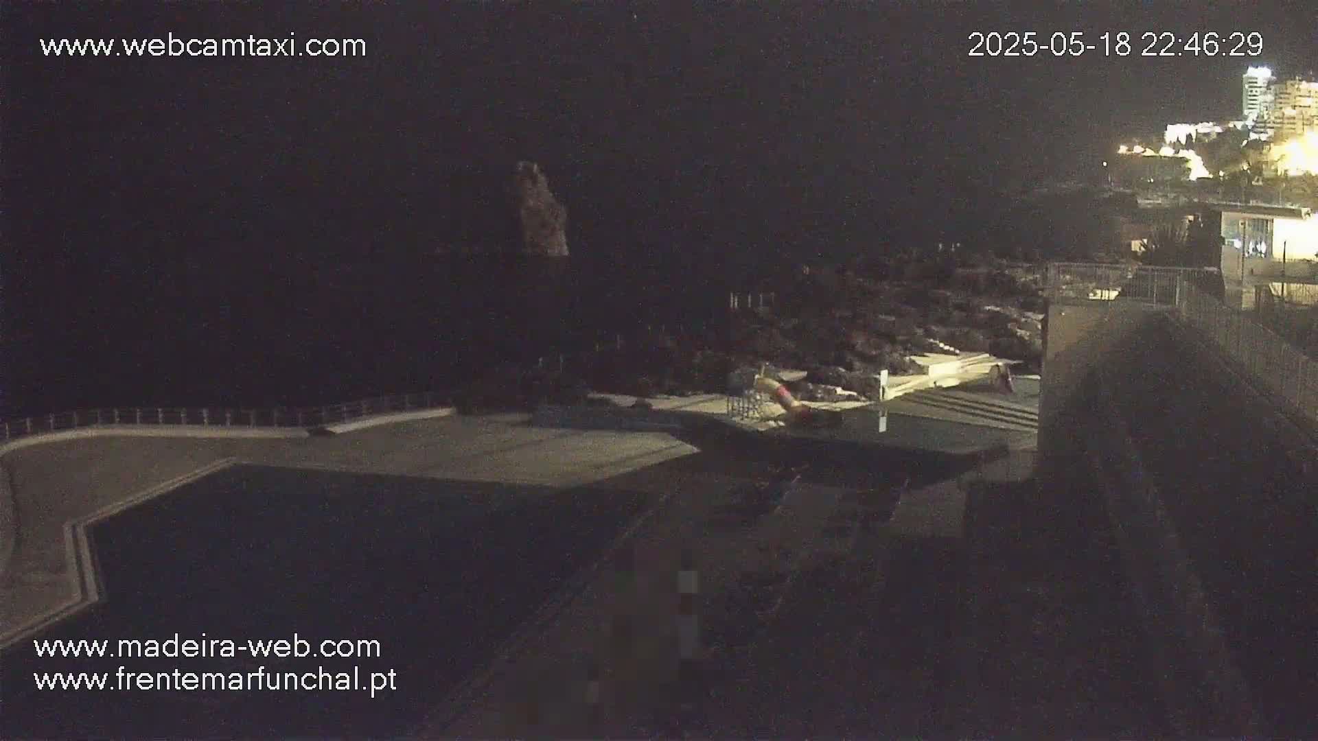 A nighttime, high-angle view of a dark, empty swimming pool next to a rocky coastline with buildings illuminated in the distance.