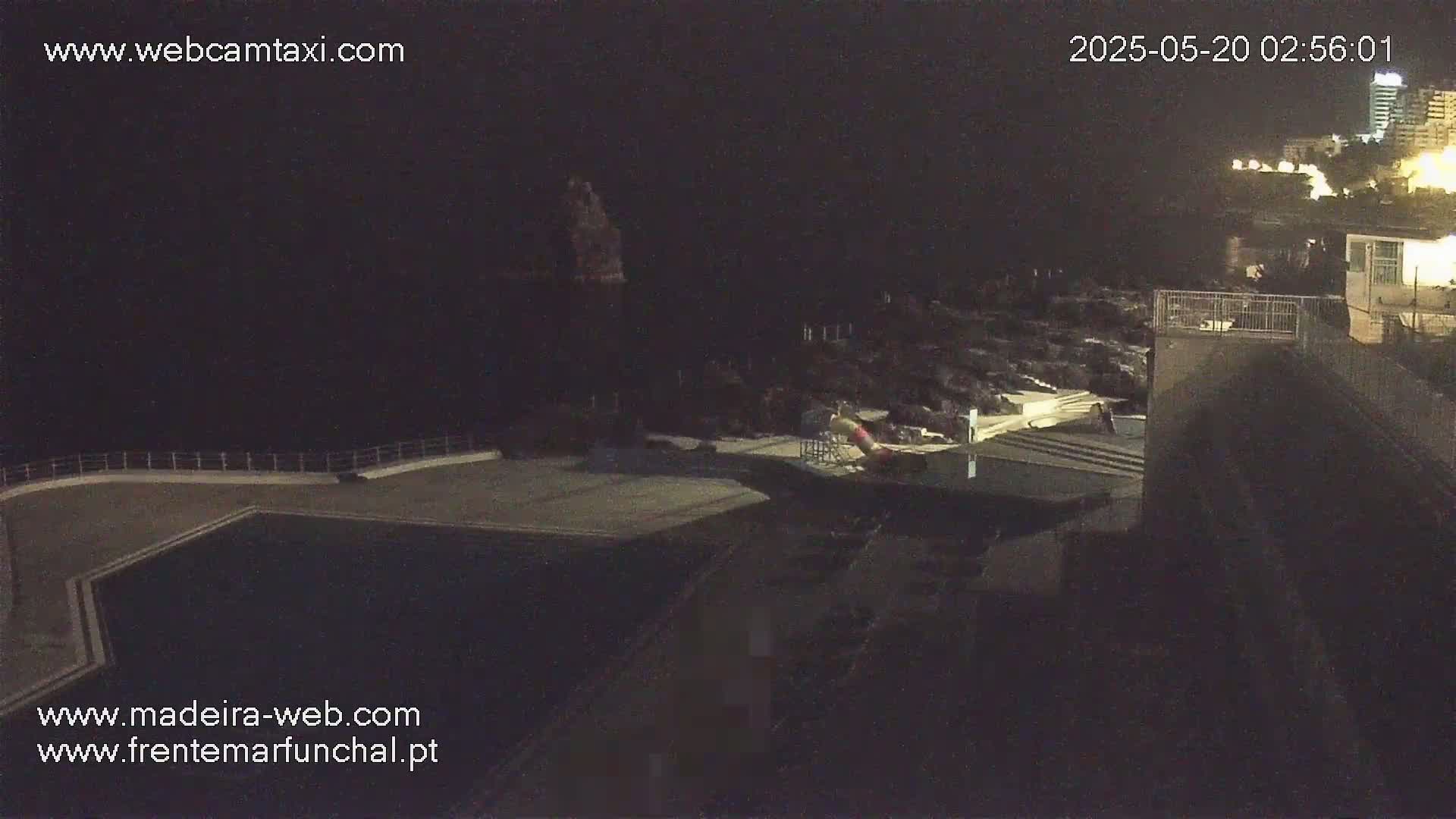 A nighttime, high-angle view shows a dark pool area next to a building overlooking a rocky coastline and distant lights.