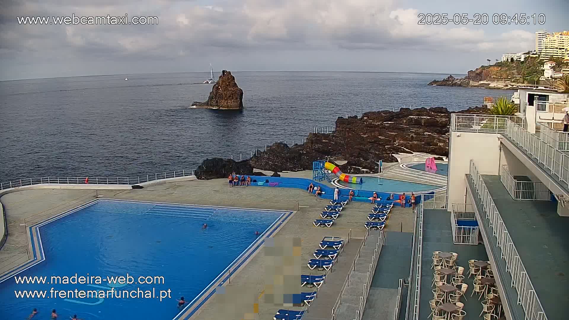 An outdoor pool complex with a large swimming pool and a smaller pool with a waterslide overlooks a rocky coastline and ocean under partly cloudy skies.
